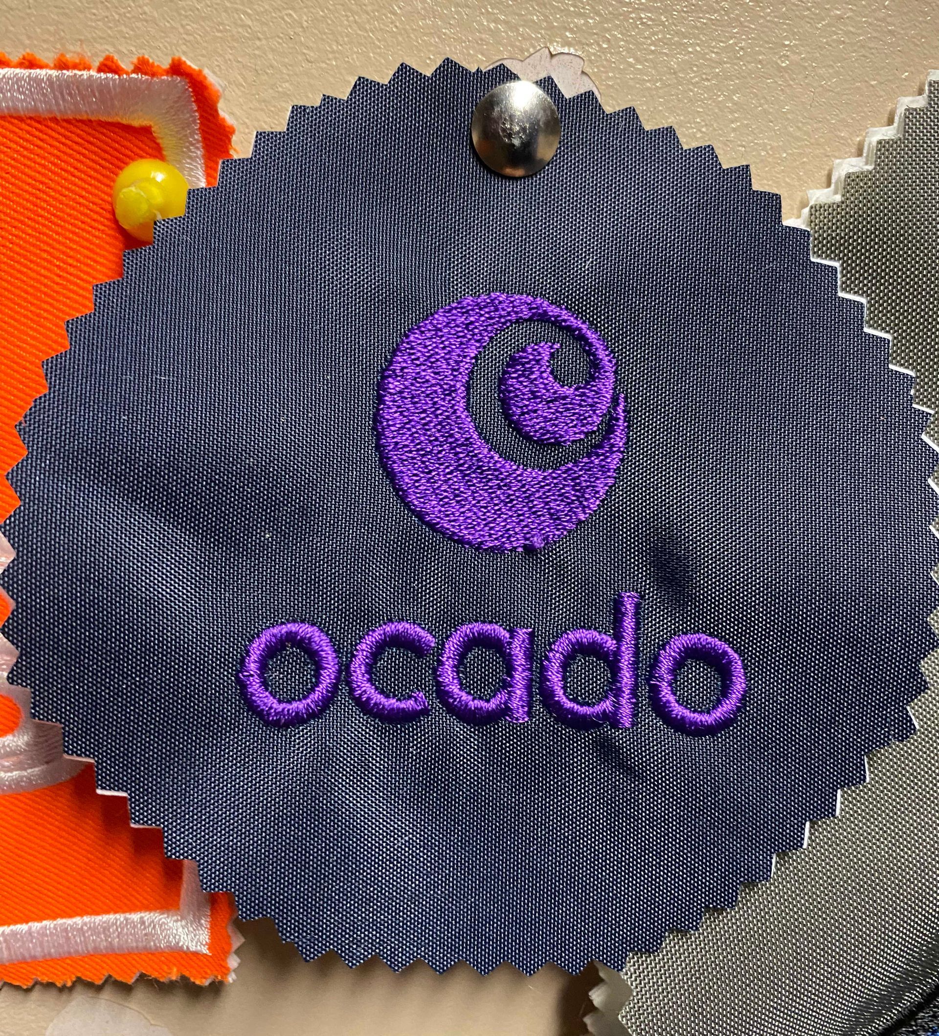 Ocado logo embroidered in purple. The swatch is dark blue with a wavy texture, pinned to a surface.