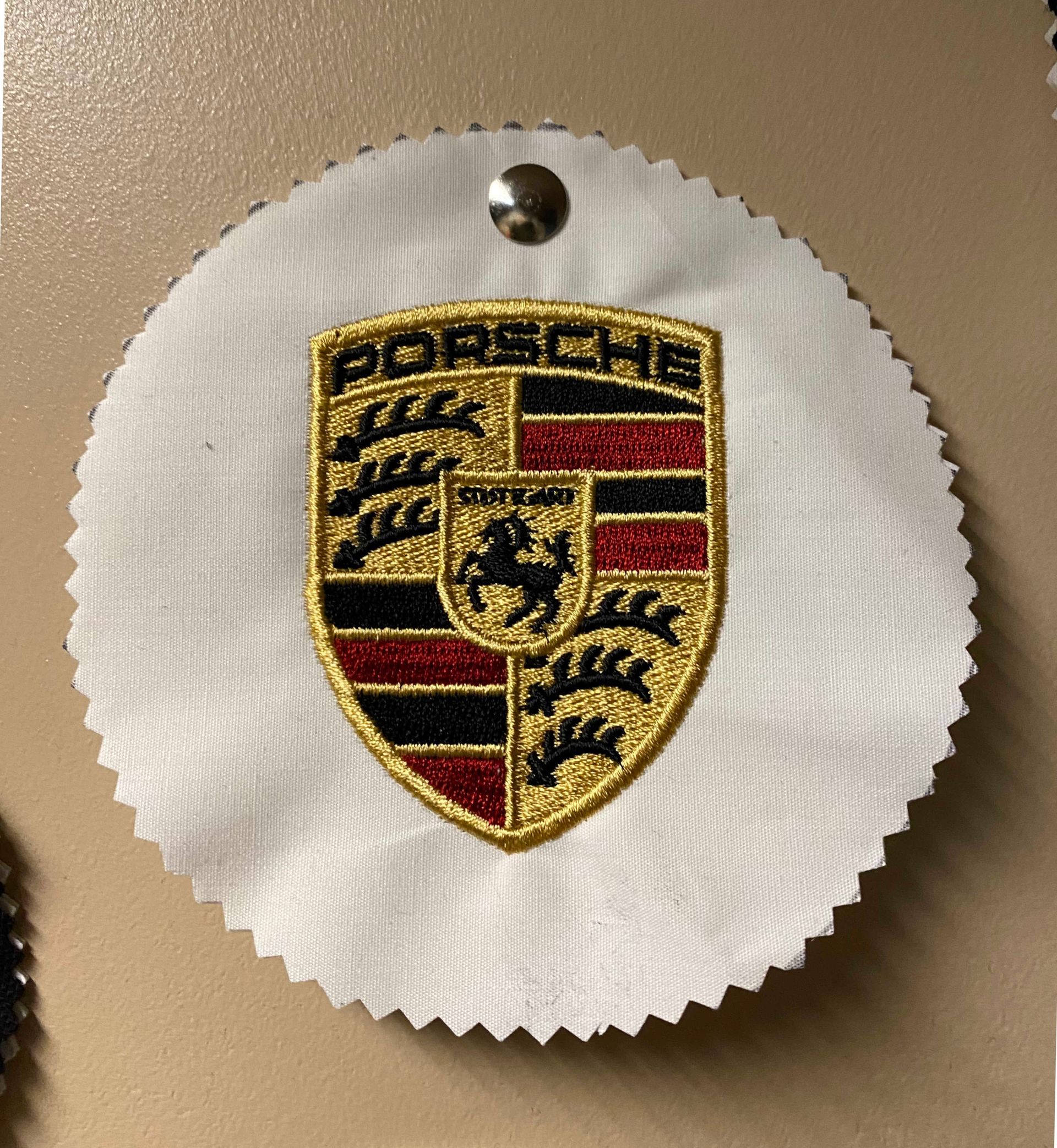 Embroidered Porsche logo on white fabric, secured with a metal tack. The logo is gold, red, black, and features a horse.