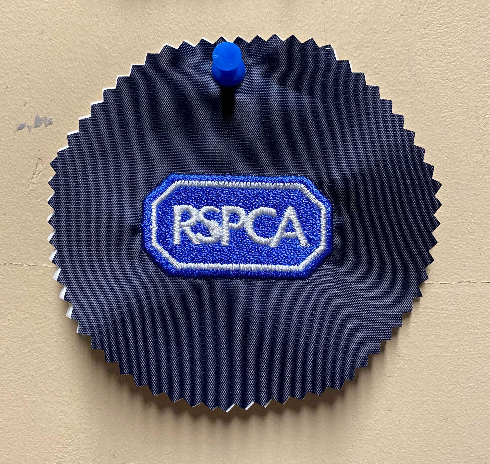 Dark blue, circular fabric swatch with a embroidered white-outlined, blue RSPCA logo pinned to a tan surface with a blue pin.