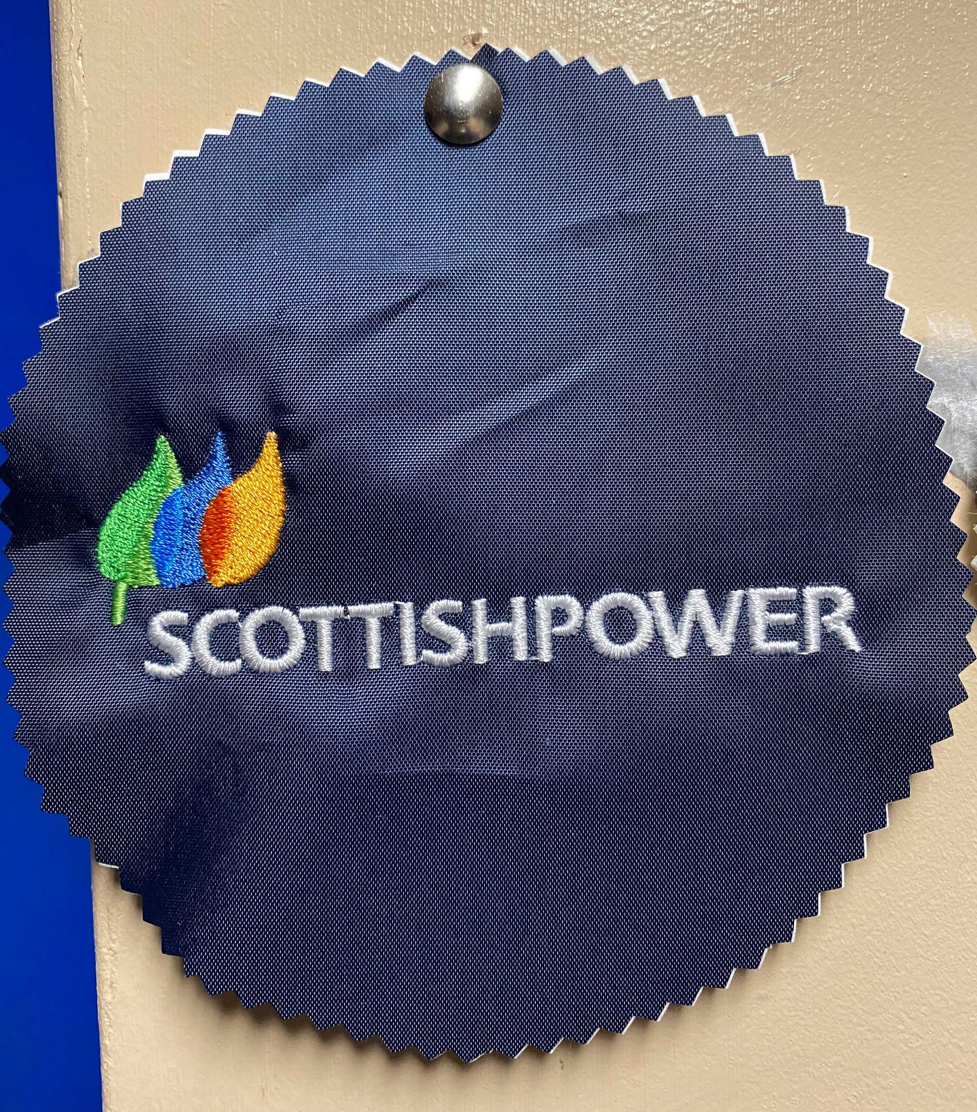 A dark blue, circular embroidered ScottishPower logo with a colorful leaf design, hanging from a wall.