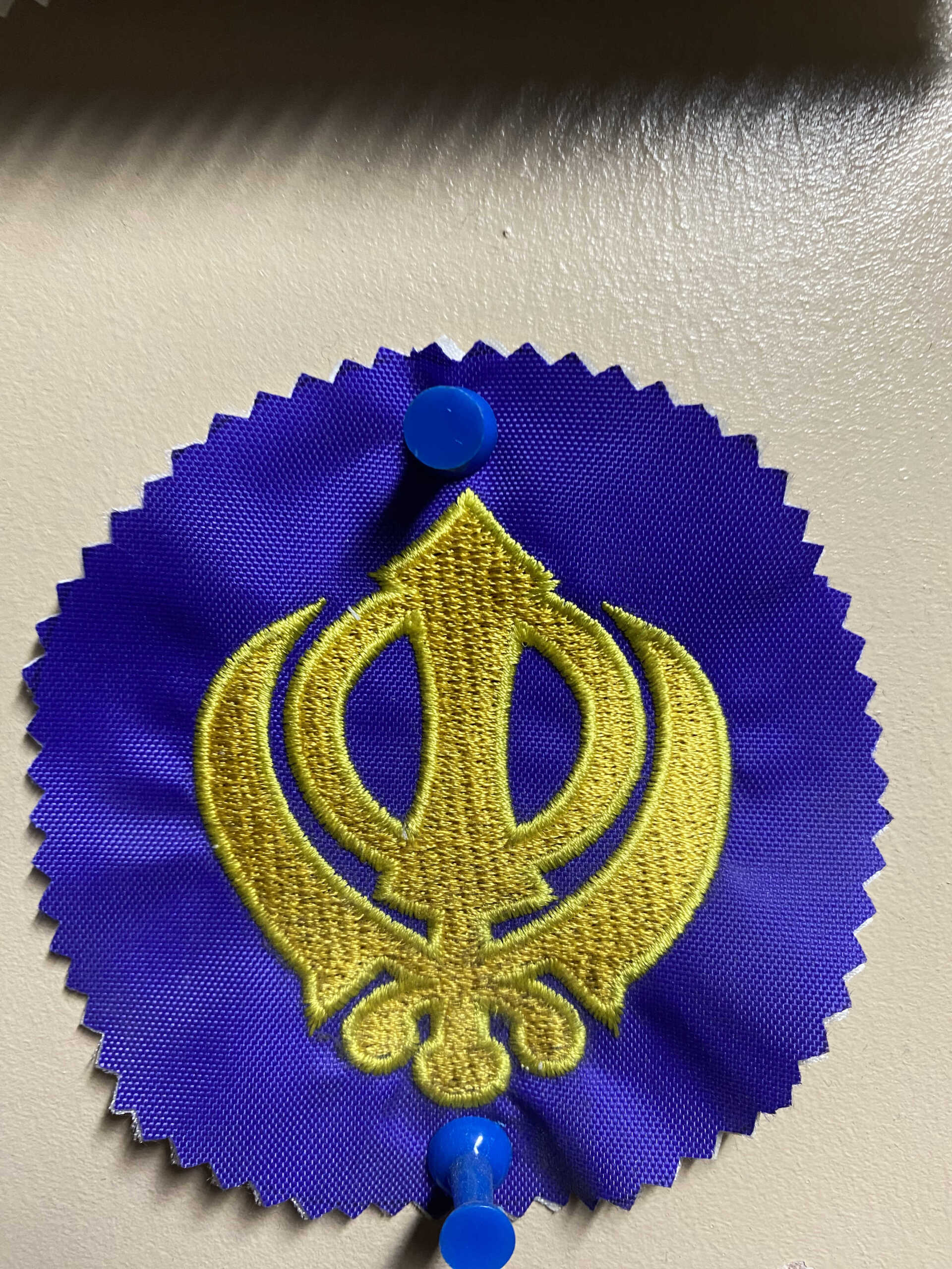 Blue rosette with gold embroidered Khanda symbol, pinned to a surface.