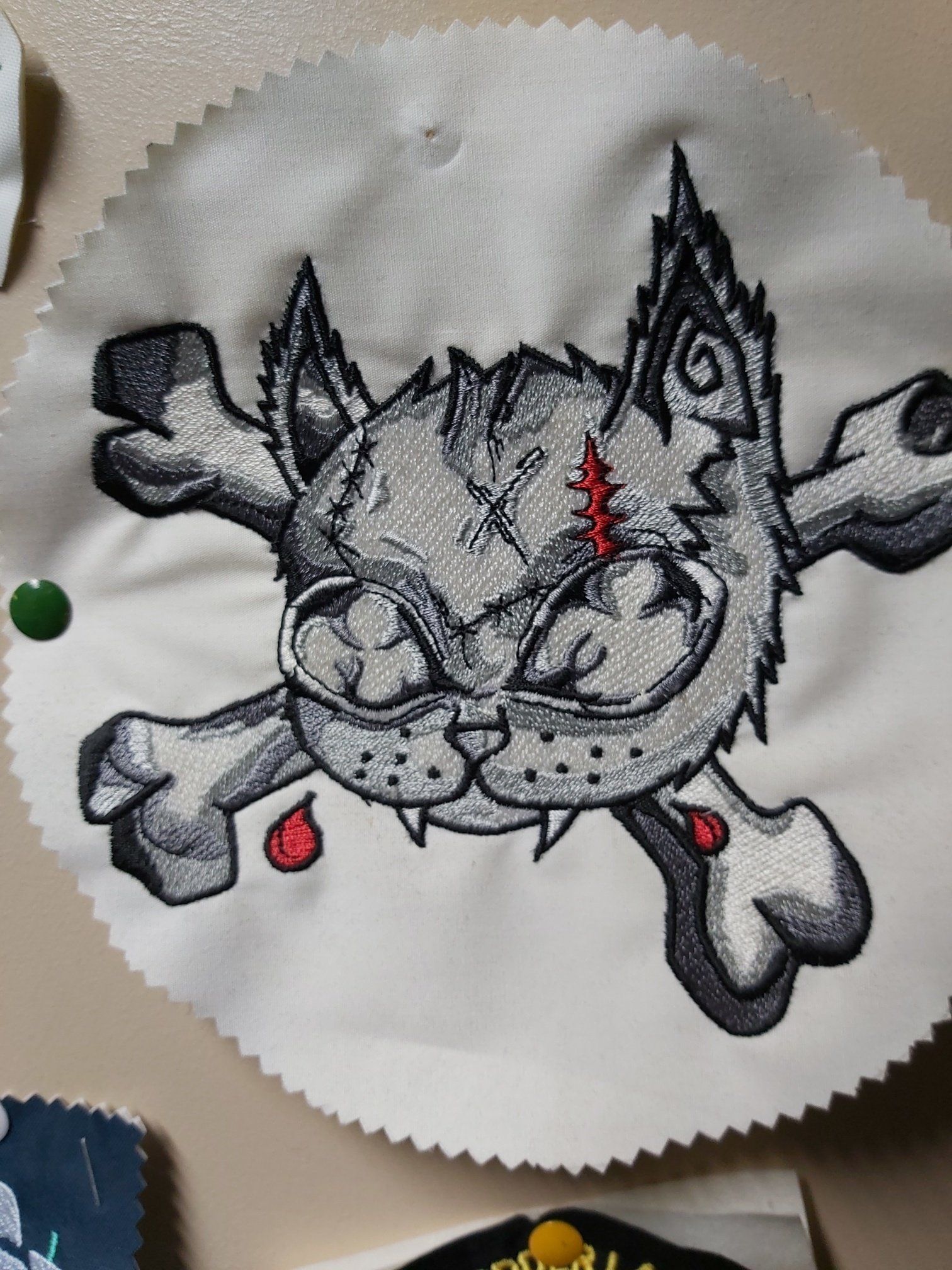 Embroidered skull with cat's face and crossed bones. Gray and white with red accents, showing a fierce expression