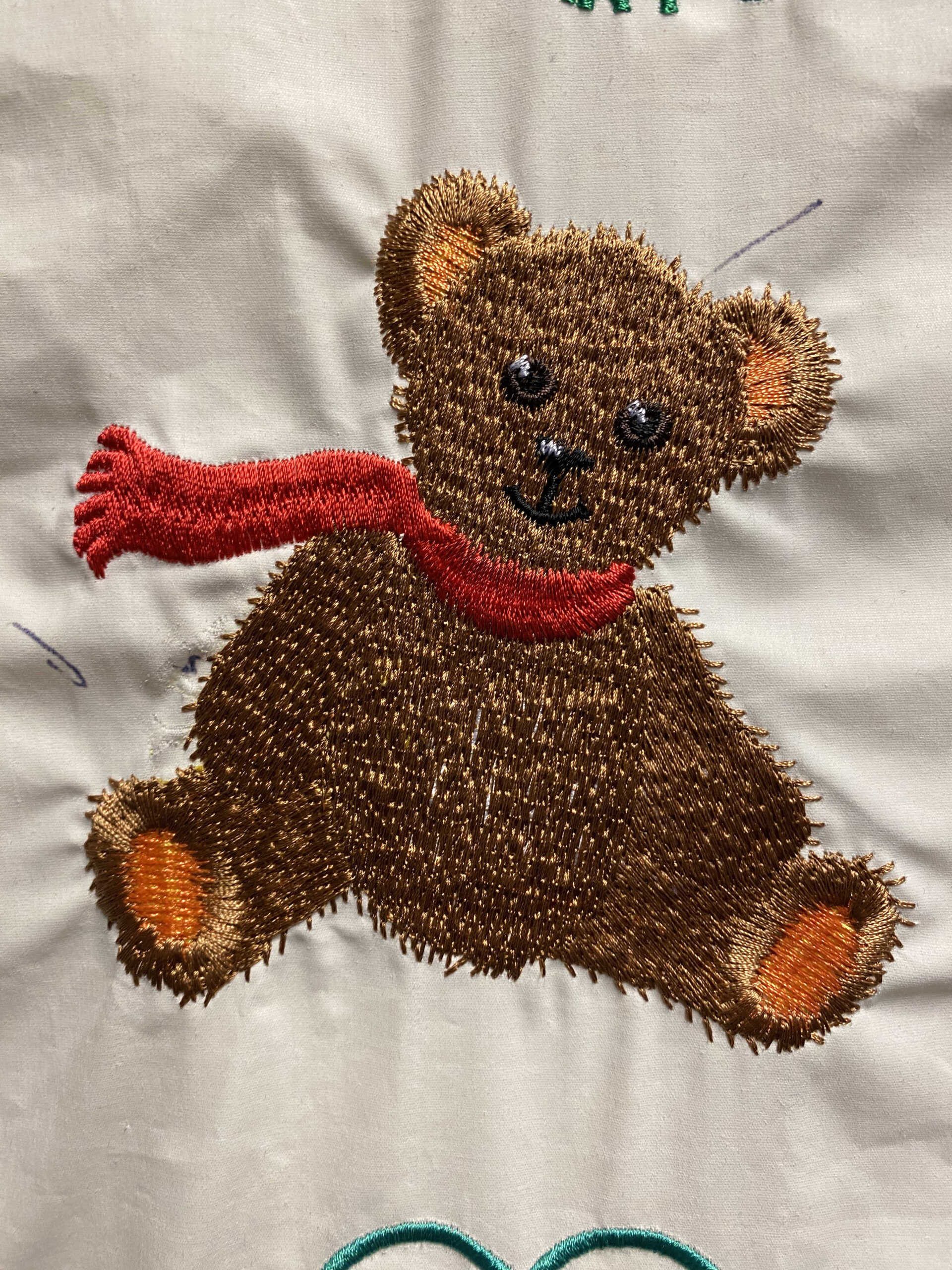 Embroidered teddy bear with brown fur and an orange scarf, sitting and smiling on cream-colored fabric.