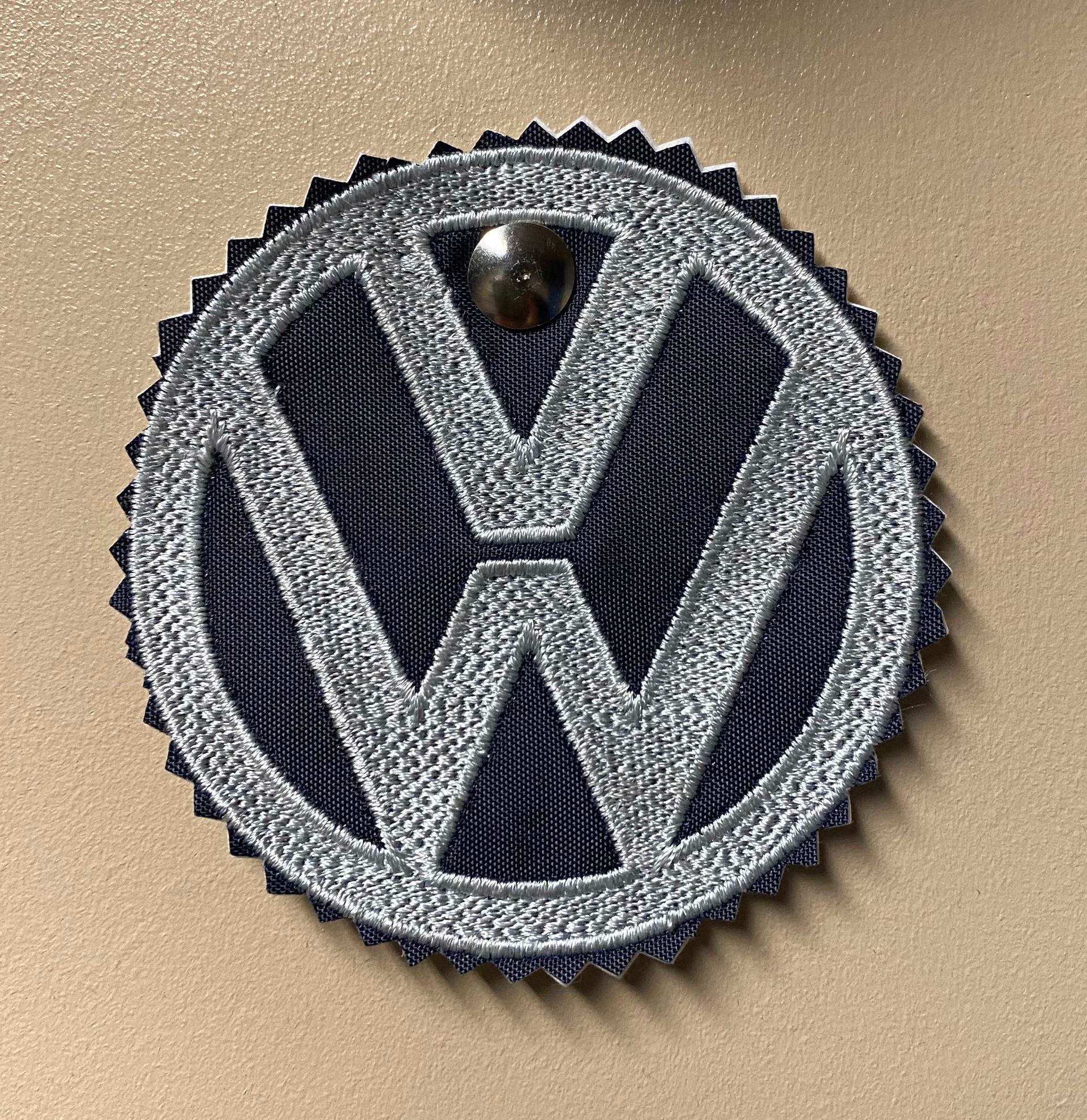 Embroidered Volkswagen logo patch with silver outline and dark blue interior, affixed to a tan surface with a single stud.