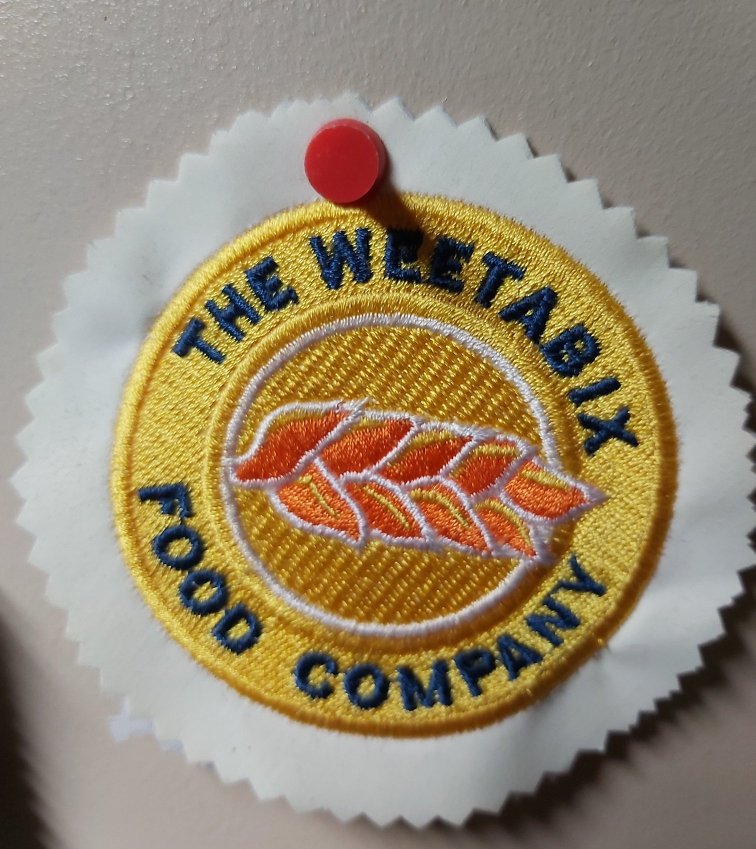 A circular yellow embroidered Weetabix food company logo, with a wheat stalk, pinned to a white background with a red pin.