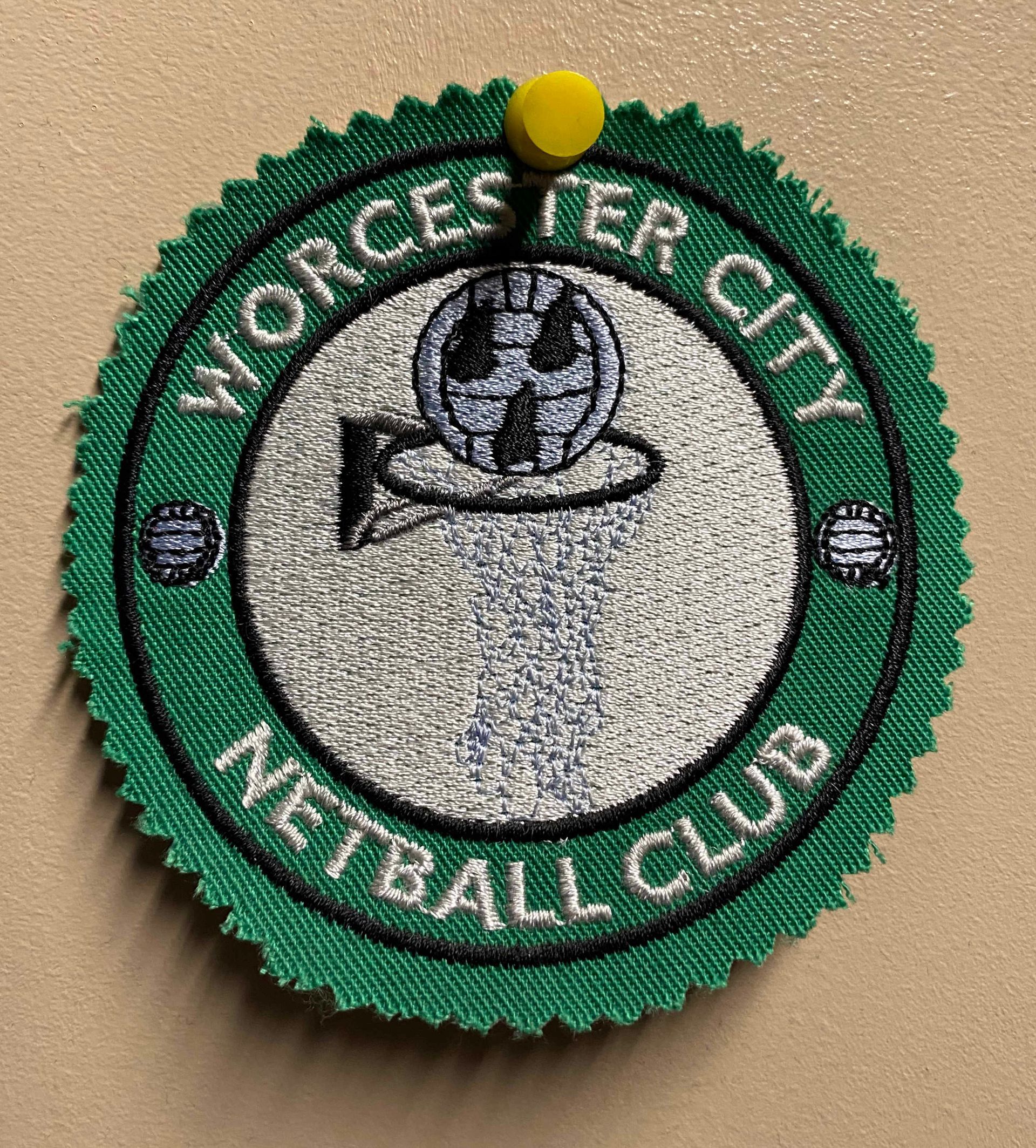 A green and white Worcester City Netball Club embroidered patch pinned to a surface. It depicts a netball within a net.
