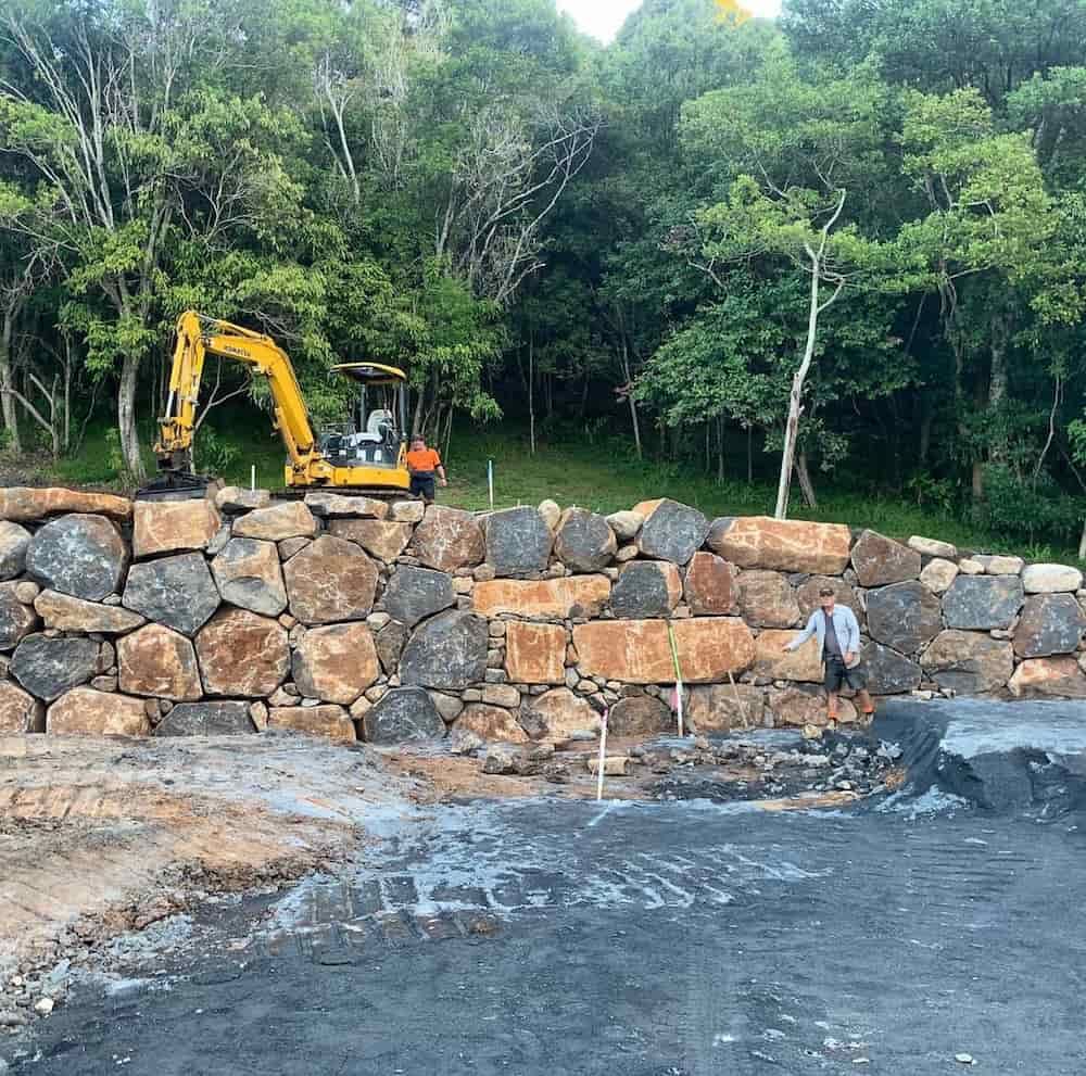 A Yellow Excavator Is Working On A Rock Wall In The Woods — J & M Bashforth & Sons In Brunswick Heads, NSW