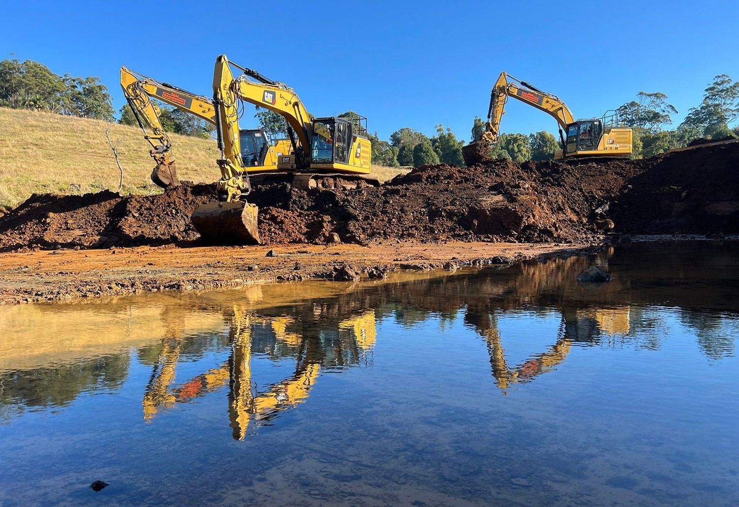 Three Yellow Excavators Working On The Edge Of A Body Of Water
