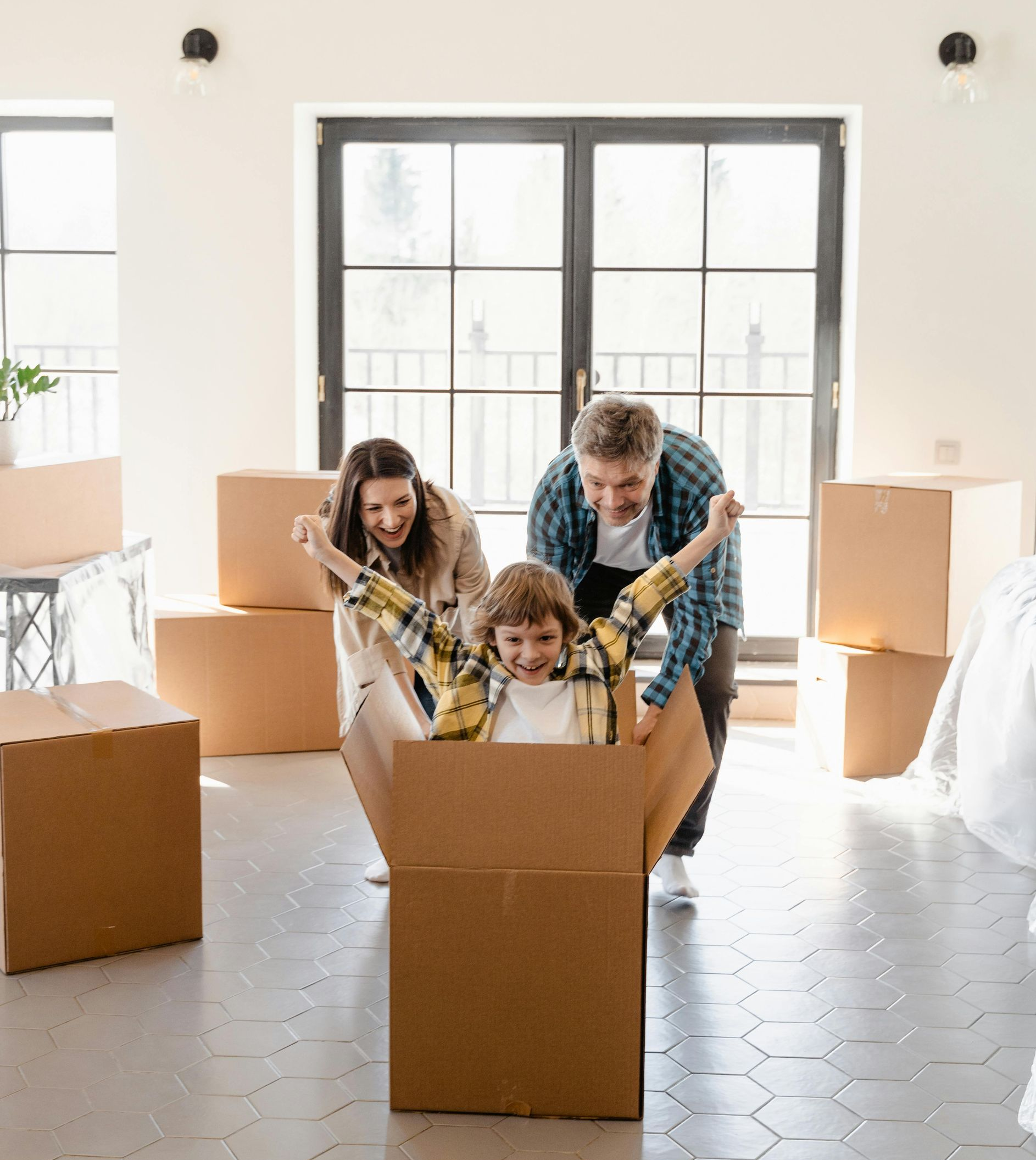 Family moving; child in cardboard box, parents pushing; light room.