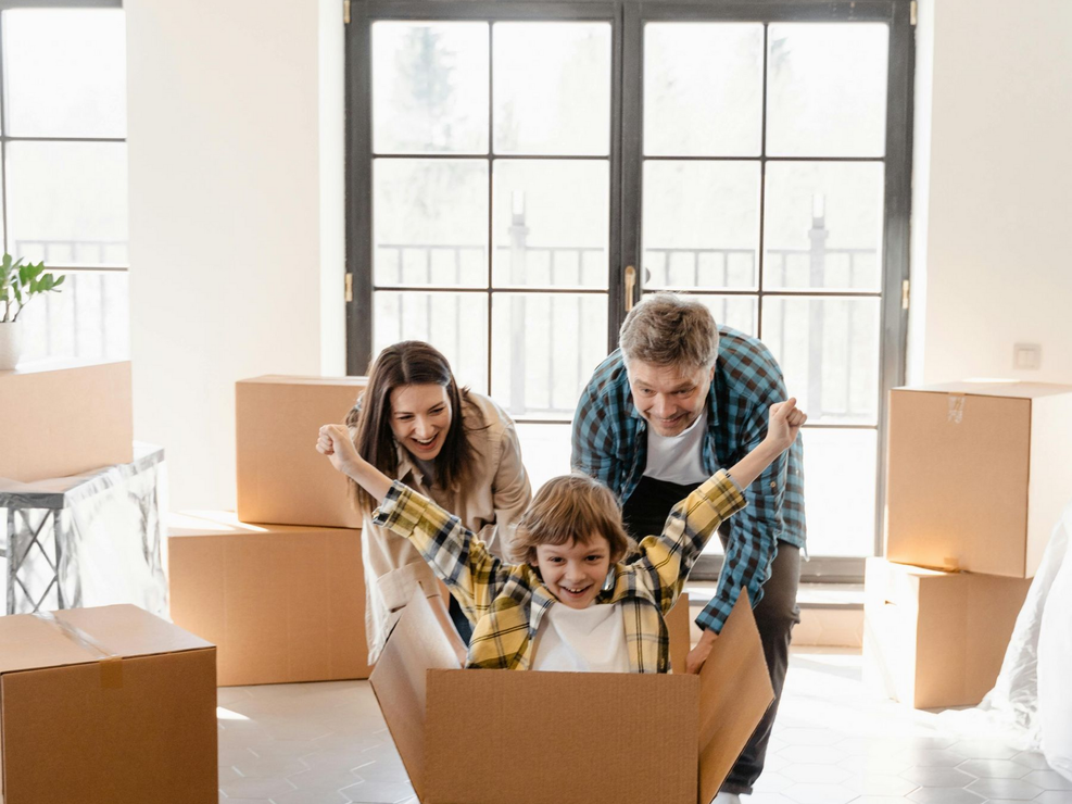 Family moving: parents pushing a child in a cardboard box, excited expressions. Boxes and windows in a bright room.