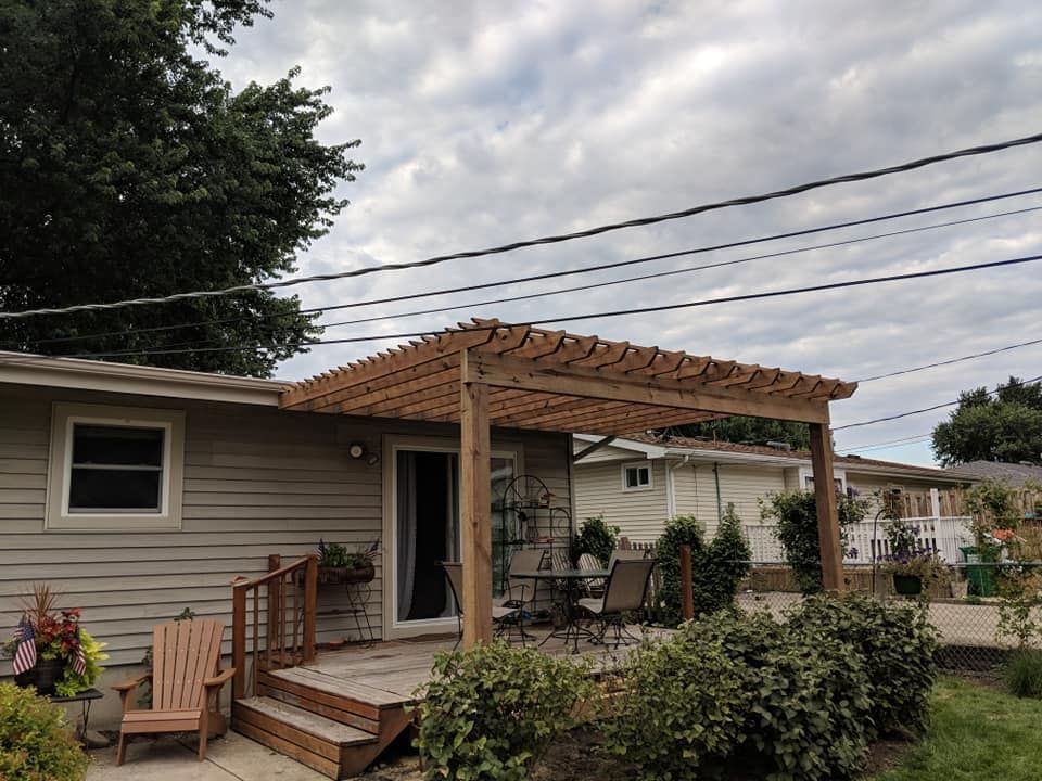 Wooden pergola over a backyard patio, with a door, furniture, and landscaping, under a cloudy sky.