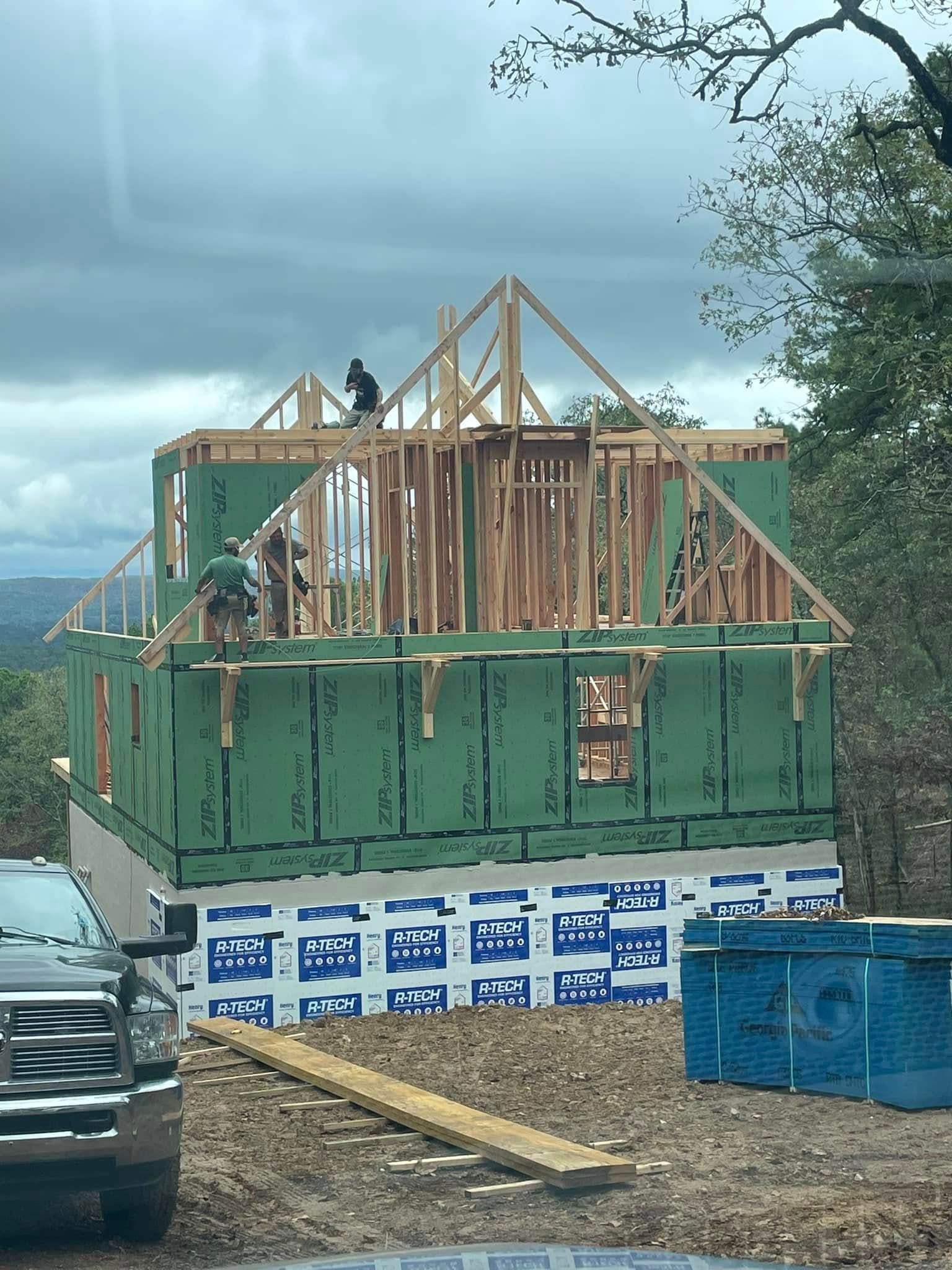Construction site: Framing a two-story house with workers. Green sheathing, blue wrap, cloudy sky.