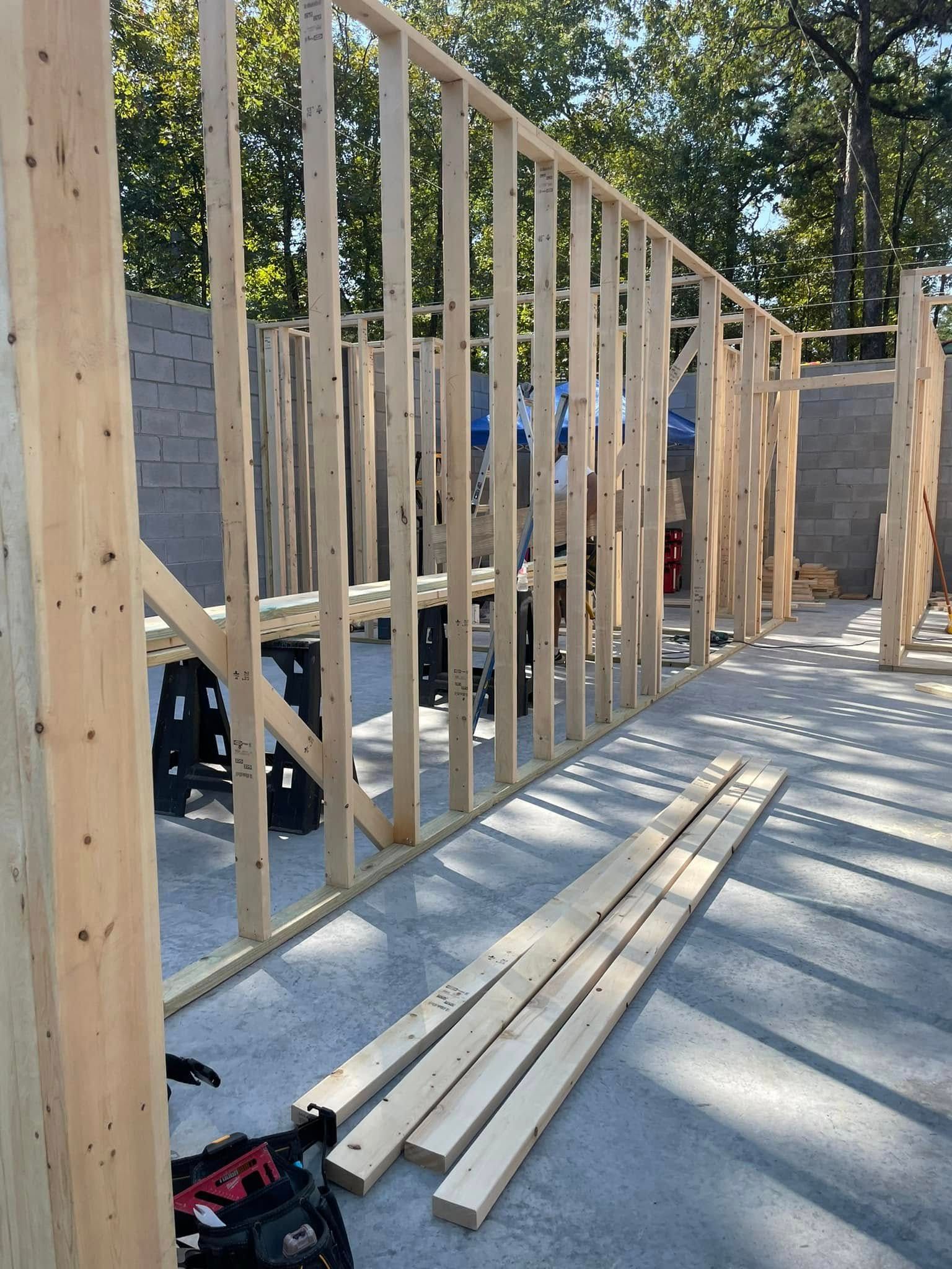Wooden studs framing a wall, set on a concrete floor.  Construction site with trees visible in the background.