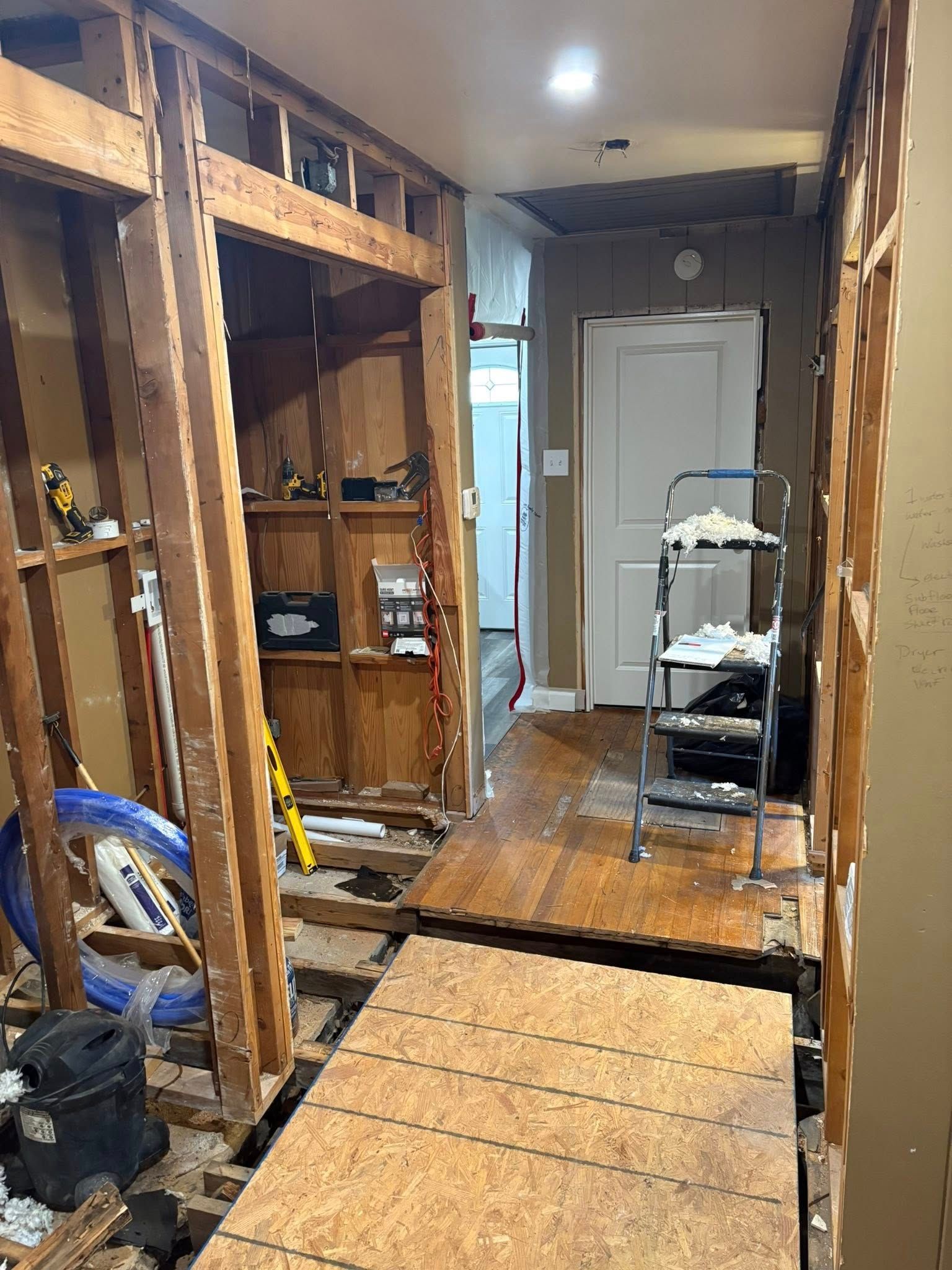 Hallway under construction with exposed wooden framing, plywood flooring, and a white door at the end.