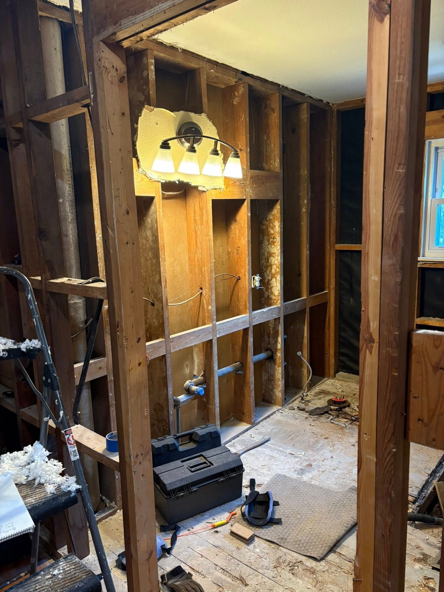 Interior of a bathroom under renovation: exposed wooden studs, plumbing, and vanity lighting.