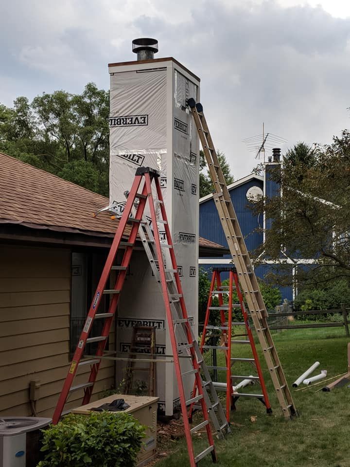 Chimney wrapped with waterproof barrier, two ladders lean against it, outdoors.