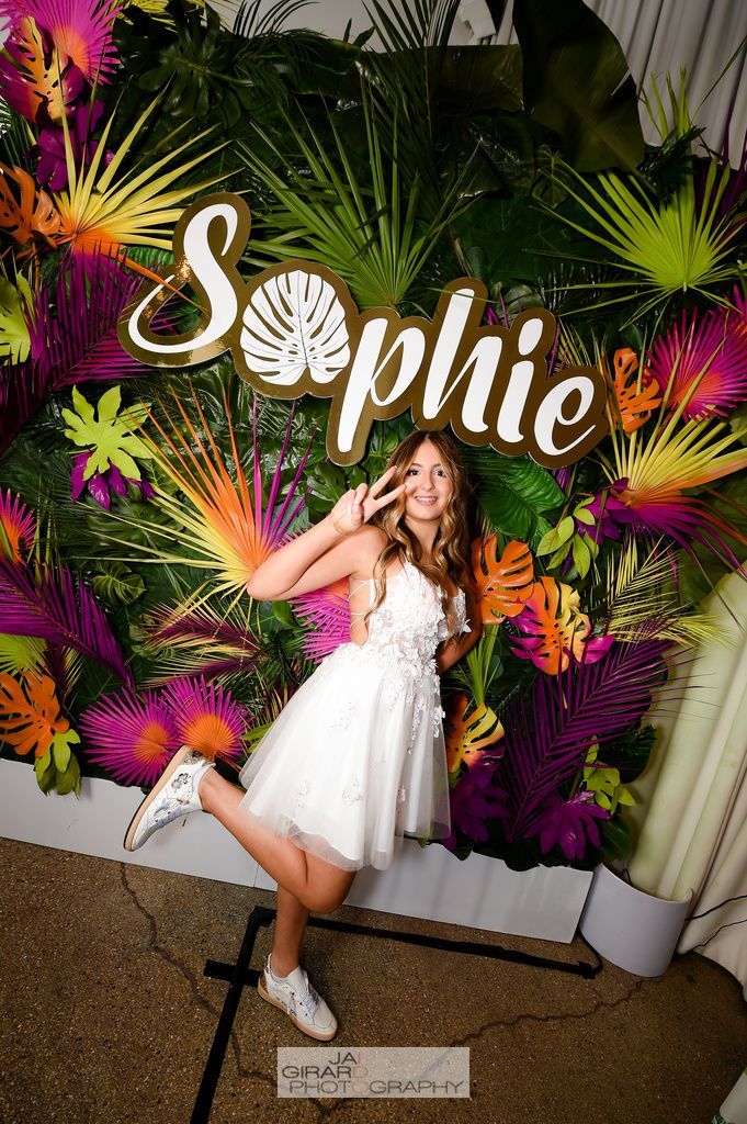 A woman in a white dress is standing in front of a wall of flowers and leaves.