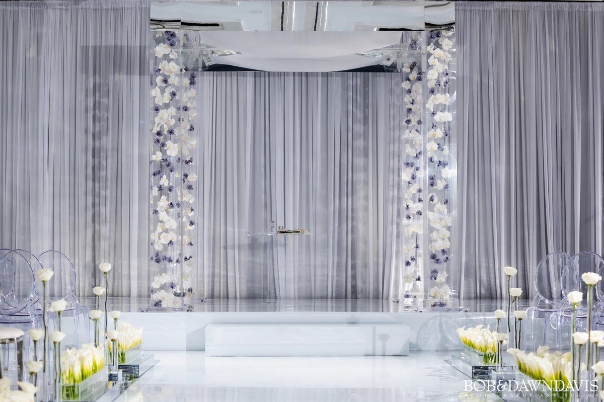 A wedding ceremony is taking place in a room with a white curtain and flowers.