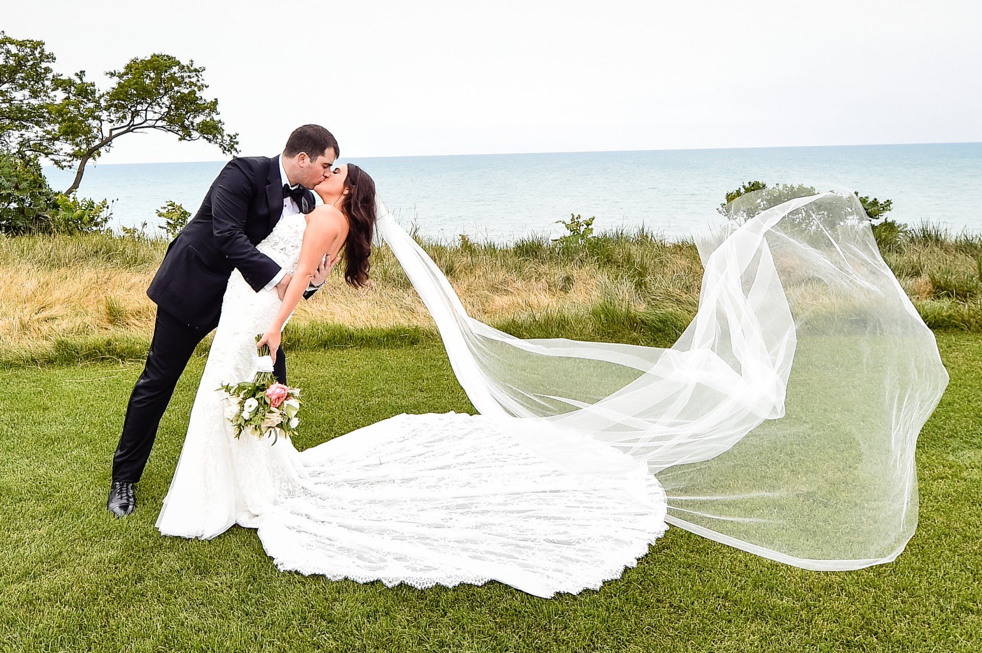 A bride and groom kissing in a field with their veil blowing in the wind.