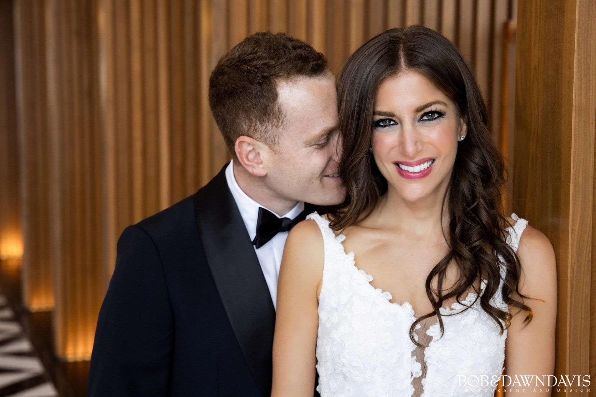 A bride and groom are posing for a picture and the groom is kissing the bride on the cheek.