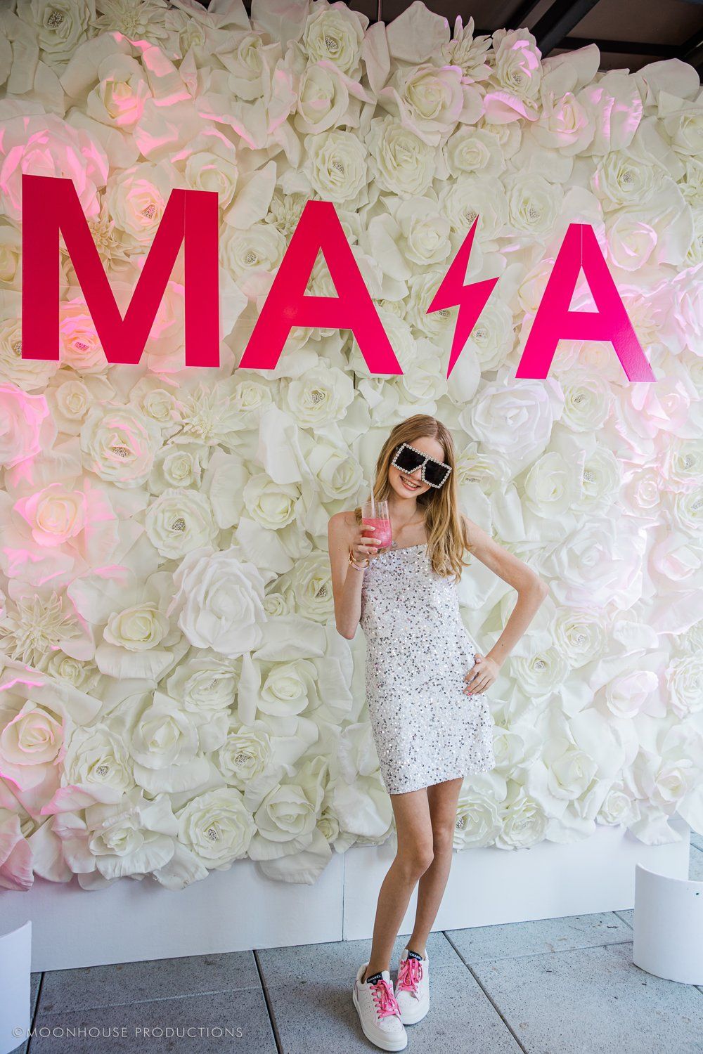 A girl in a white dress is standing in front of a wall of white flowers.