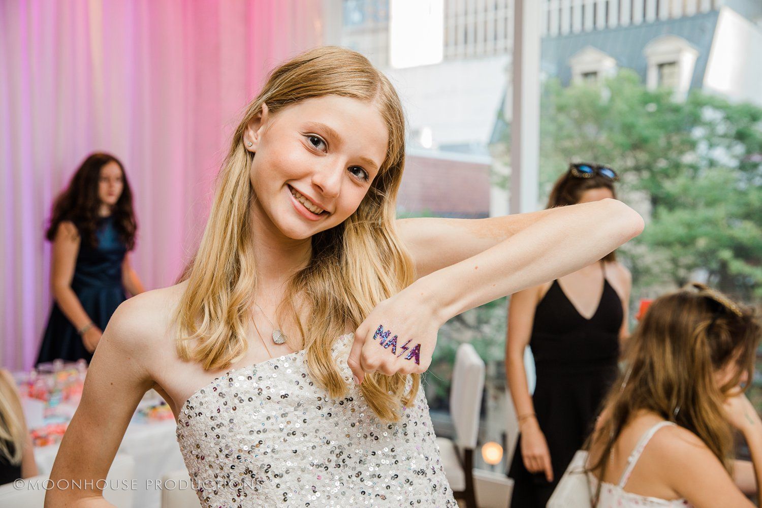 A young girl in a white dress is holding her arm up and smiling.