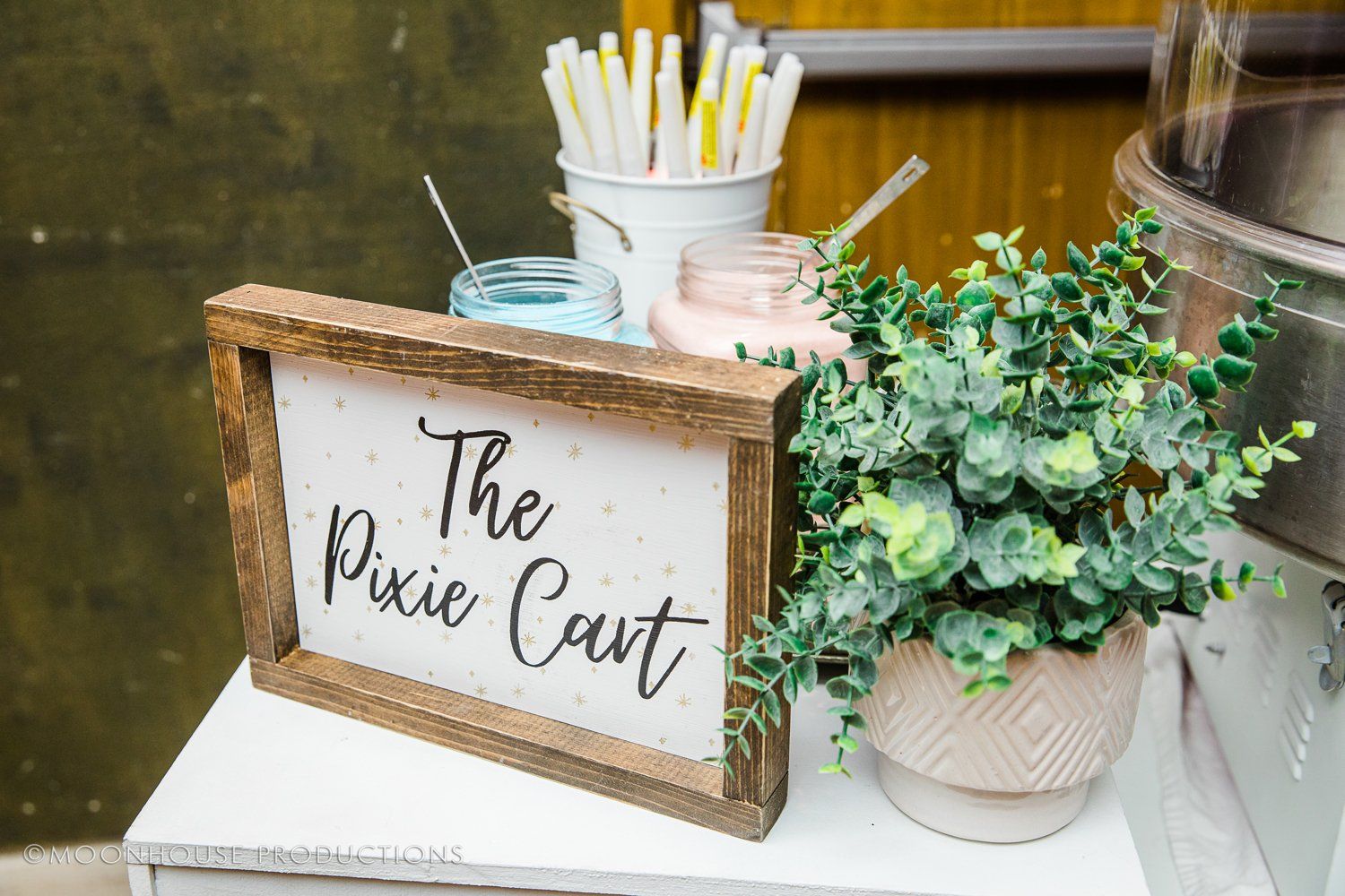 A sign that says `` the pixie cart '' is sitting on a table next to a potted plant.