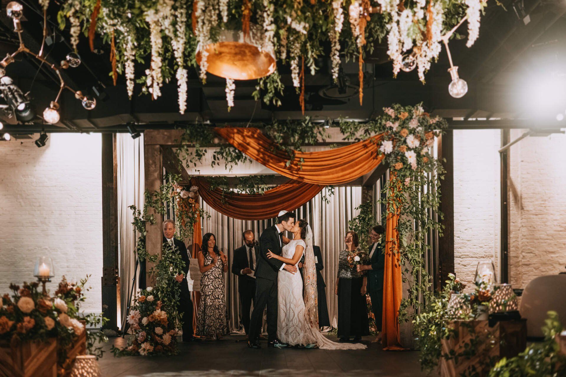 A bride and groom are kissing during their wedding ceremony in a room decorated with flowers.