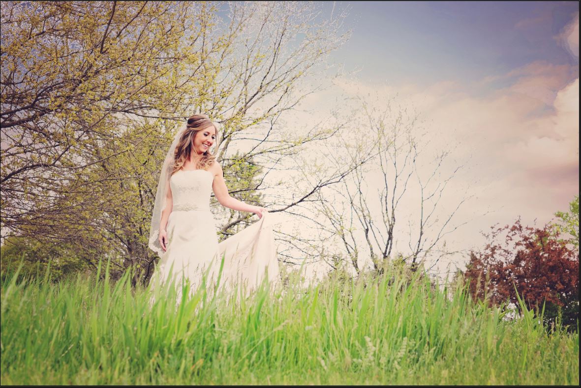 A bride in a white dress and veil is standing in a grassy field.
