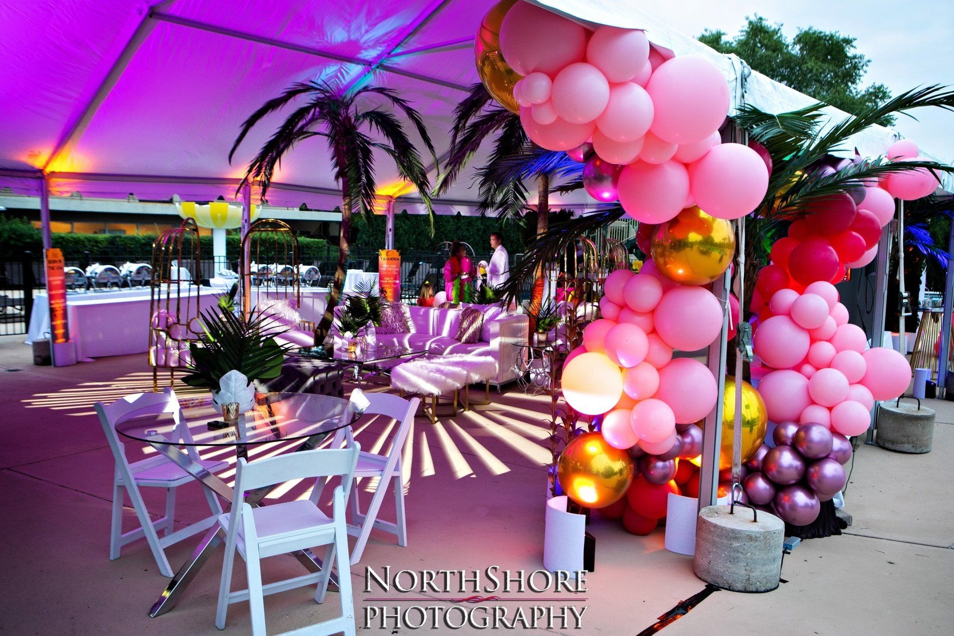 A tent is decorated with pink and gold balloons and palm trees.