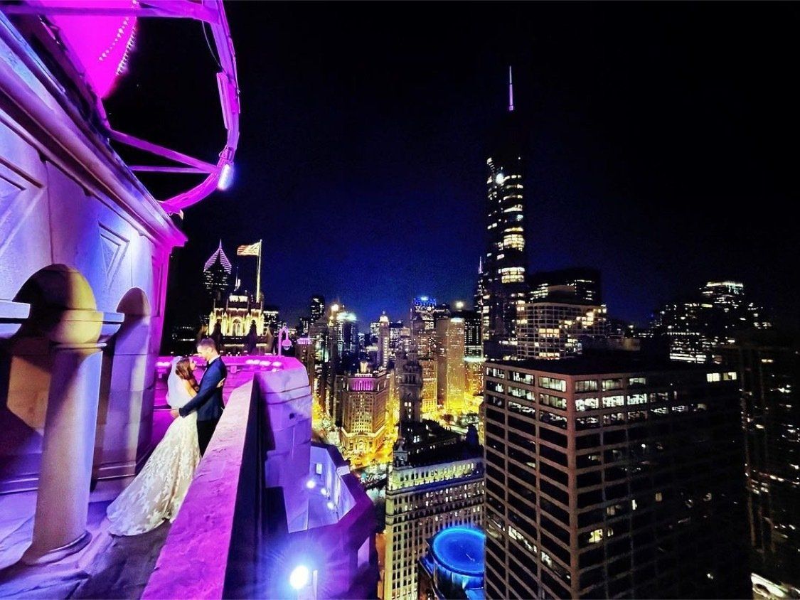 A bride and groom are standing on a balcony overlooking a city at night