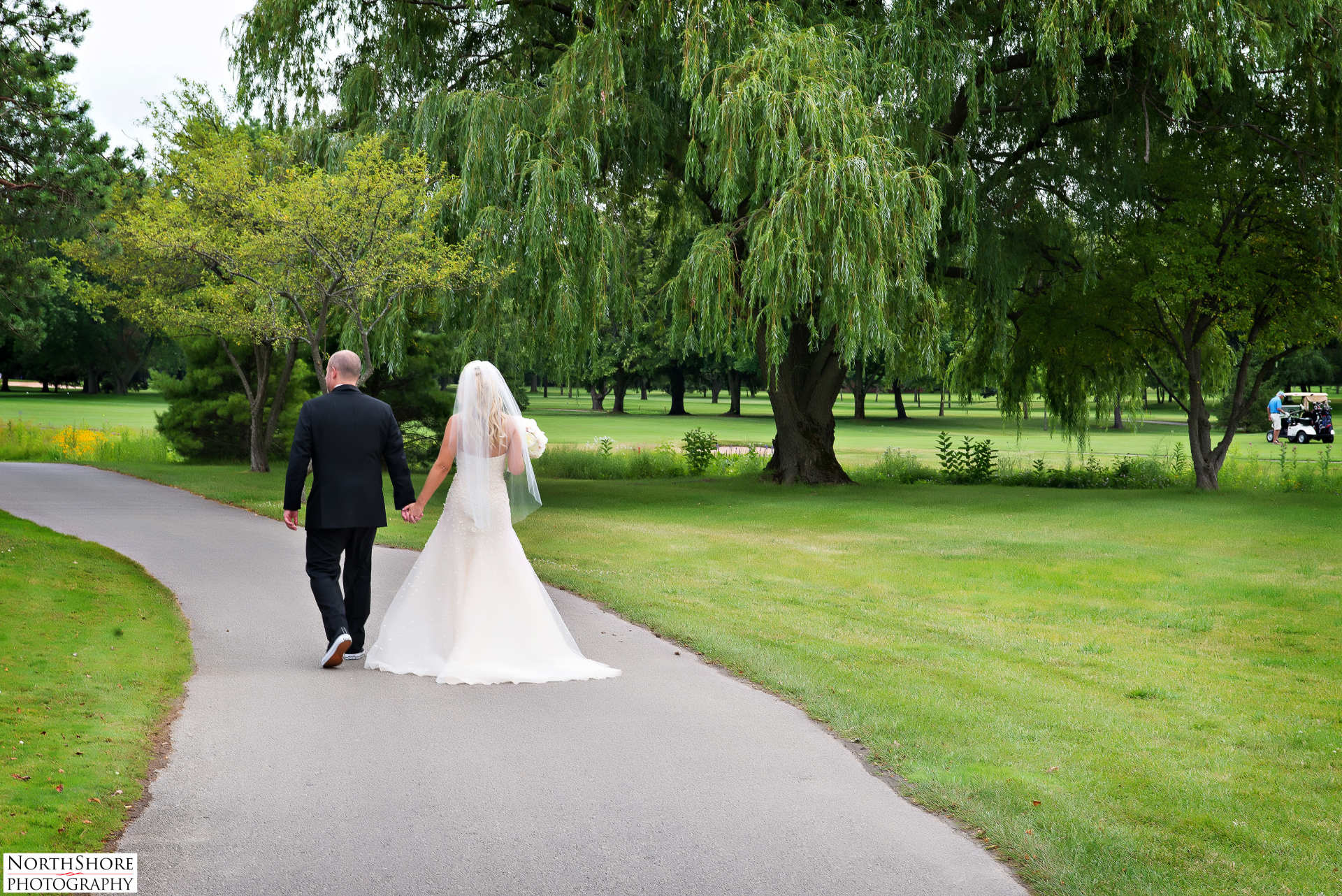 A bride and groom are walking down a path in a park holding hands.