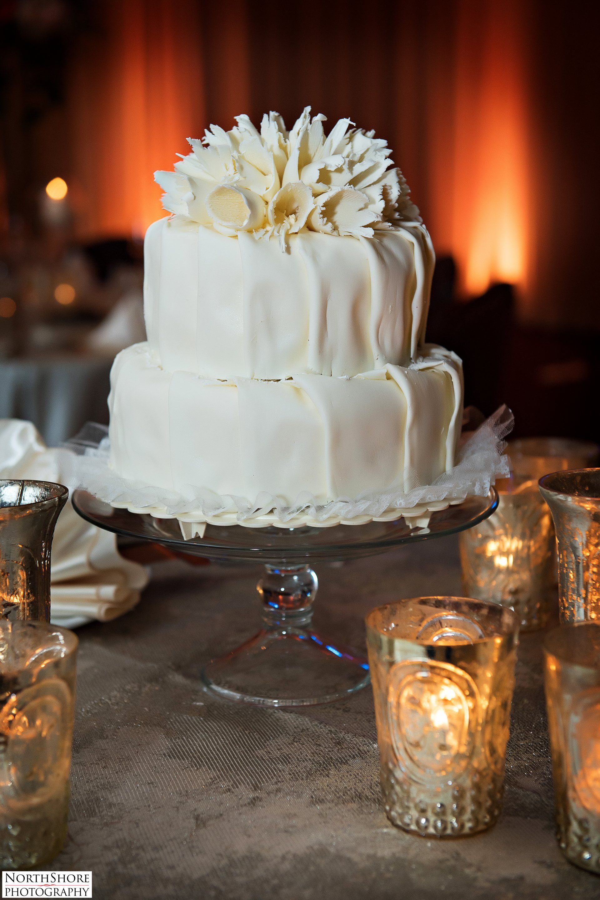 A wedding cake is sitting on a glass plate on a table surrounded by candles.