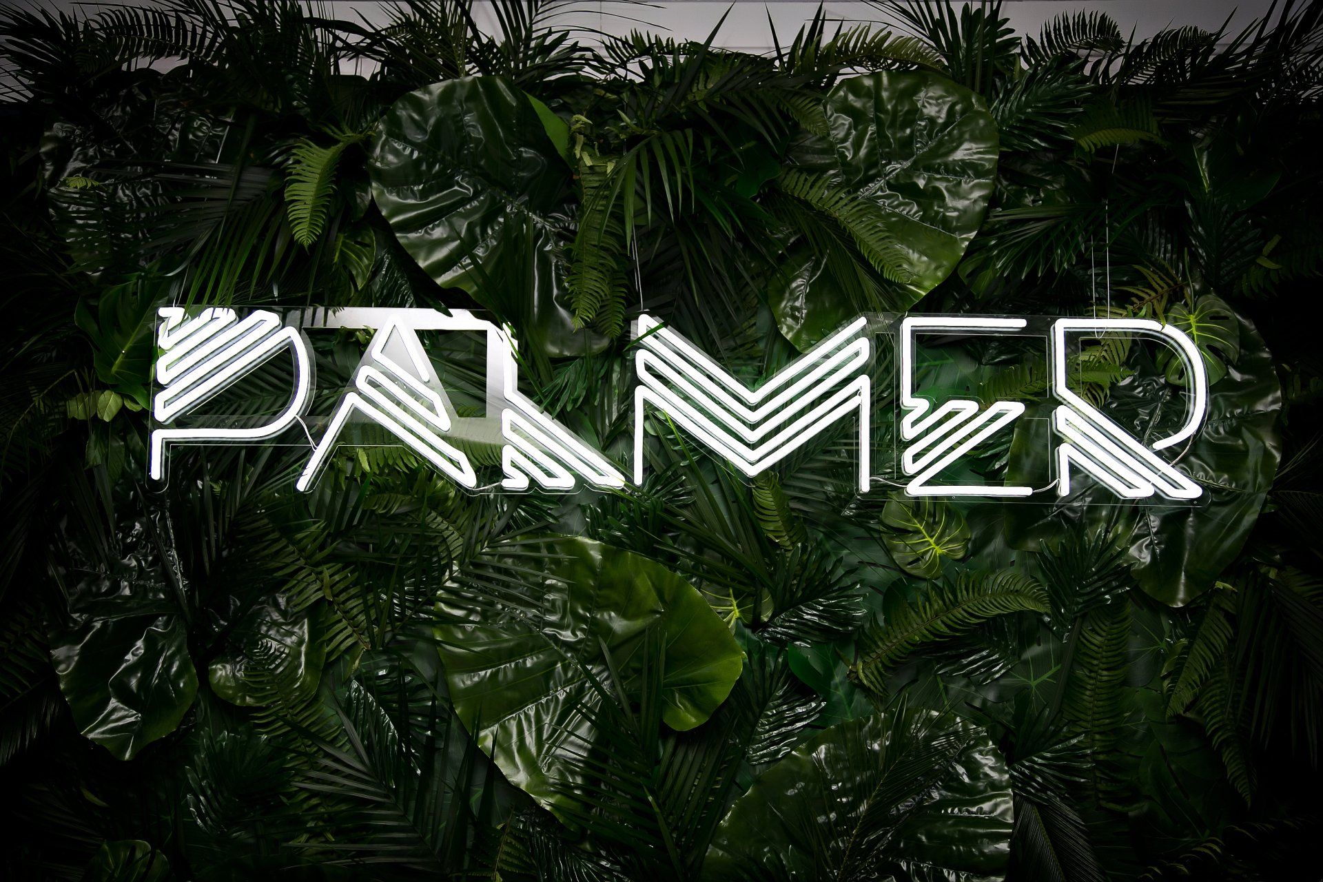 A neon sign that says parmer is hanging on a wall surrounded by green leaves.