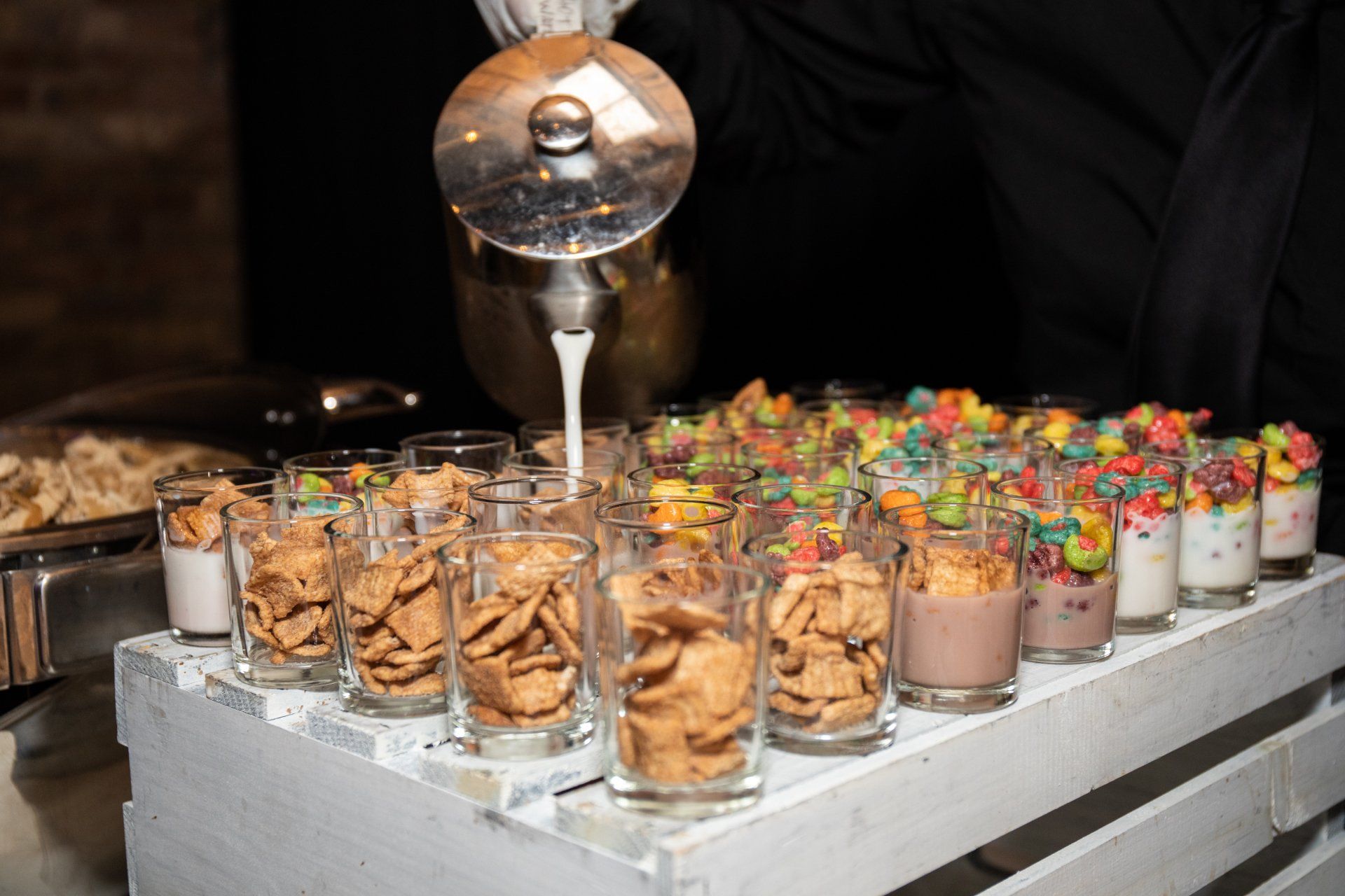 A person is pouring milk into shot glasses filled with cereal and yogurt.