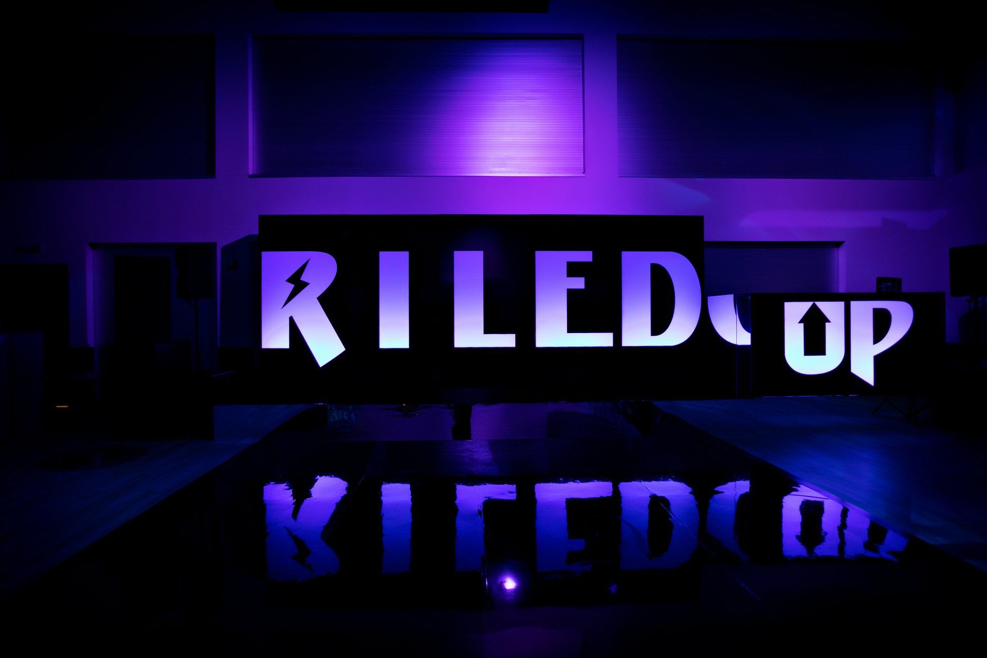 A sign that says riled up is lit up with purple lights