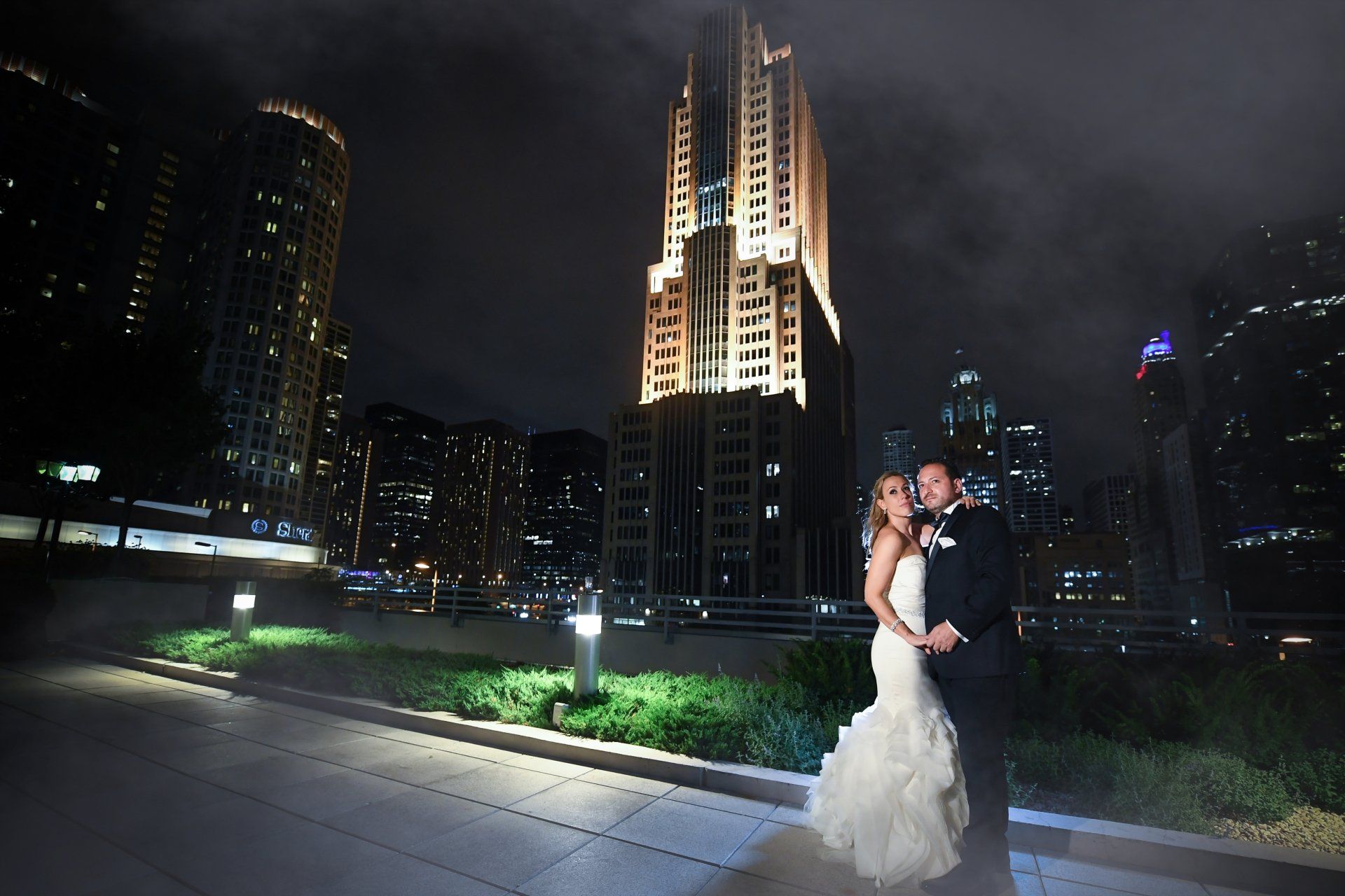 A bride and groom pose in front of a tall building at night