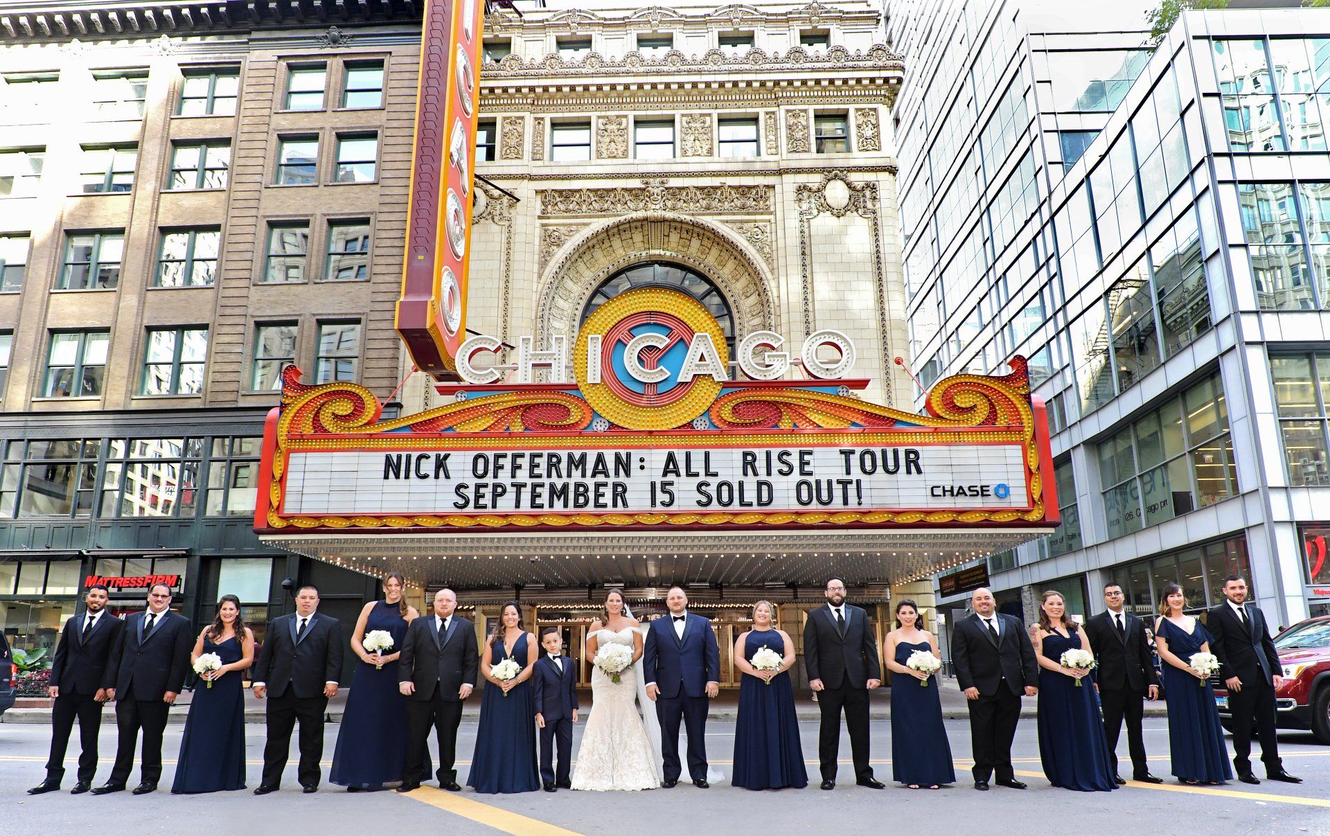 A wedding party is posing for a picture in front of a theater.