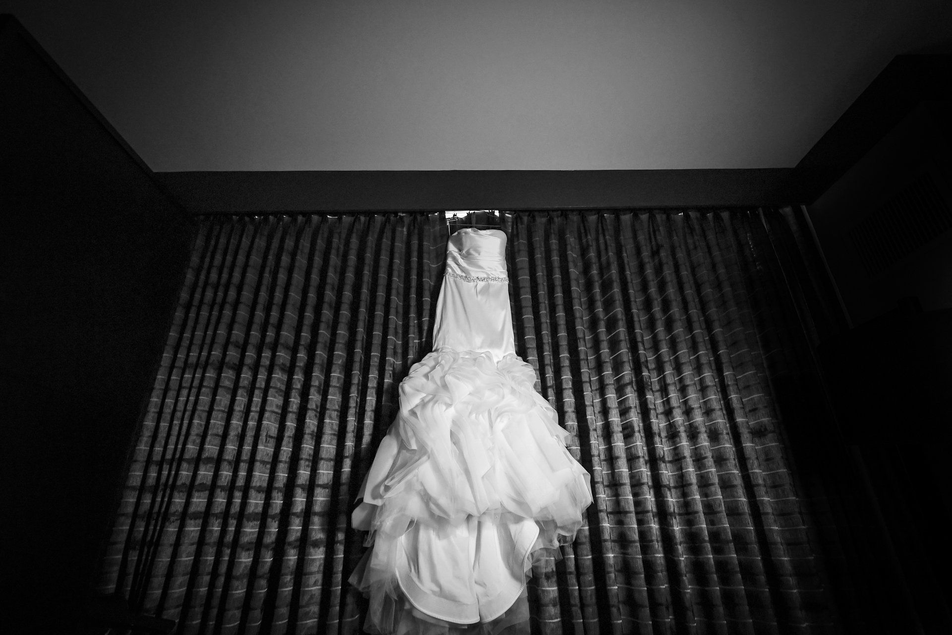 A black and white photo of a wedding dress hanging on a curtain.