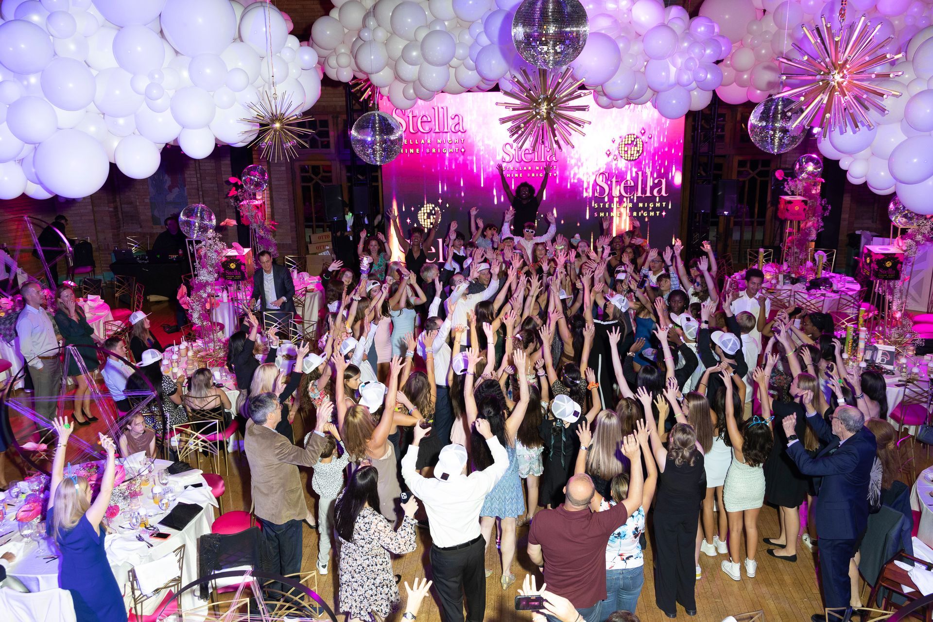 A large group of people are dancing at a party with balloons hanging from the ceiling.