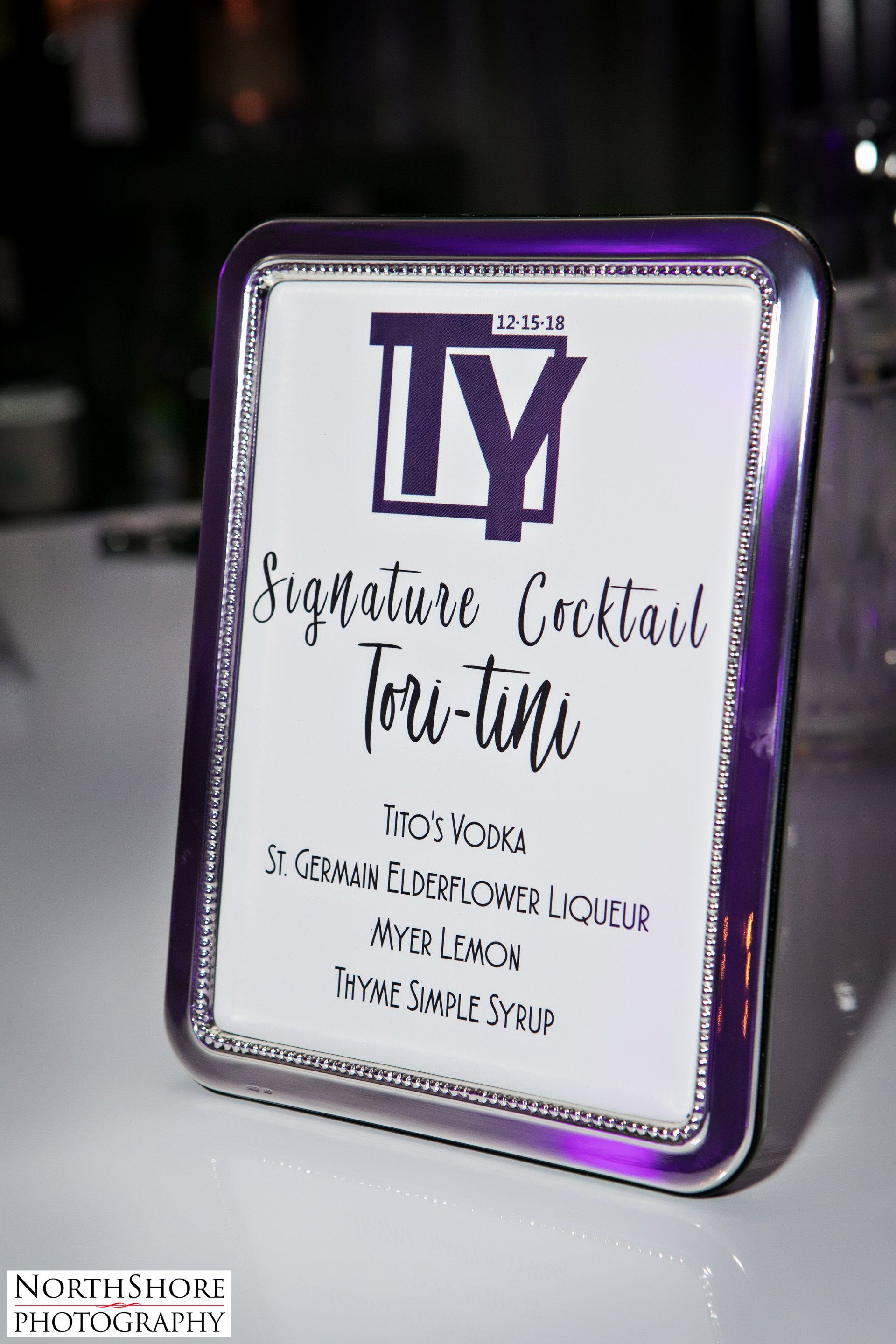 A purple and white sign that says `` signature cocktail tori-tini '' is sitting on a table.