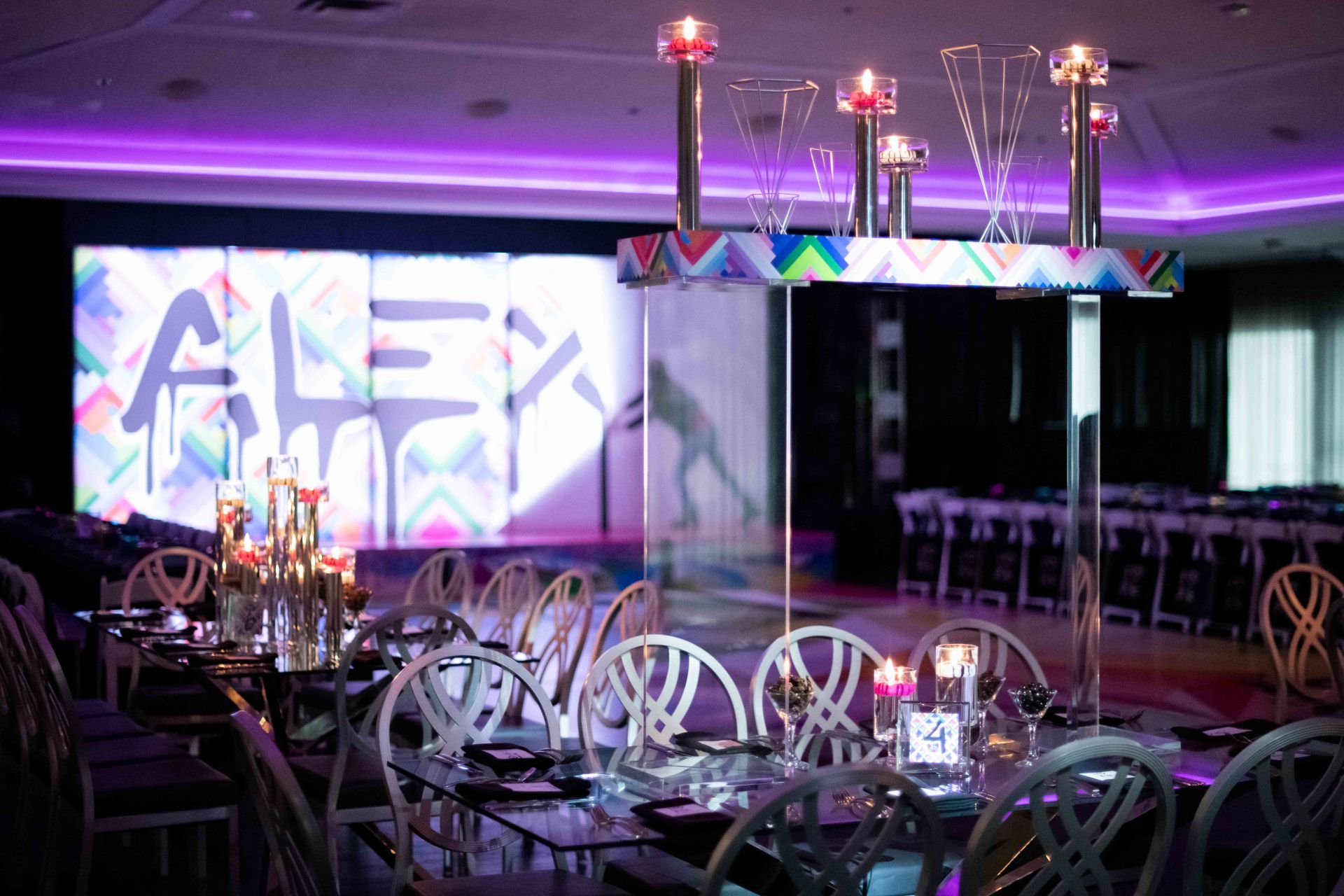 A room with tables and chairs and a large screen with the name alex on it.