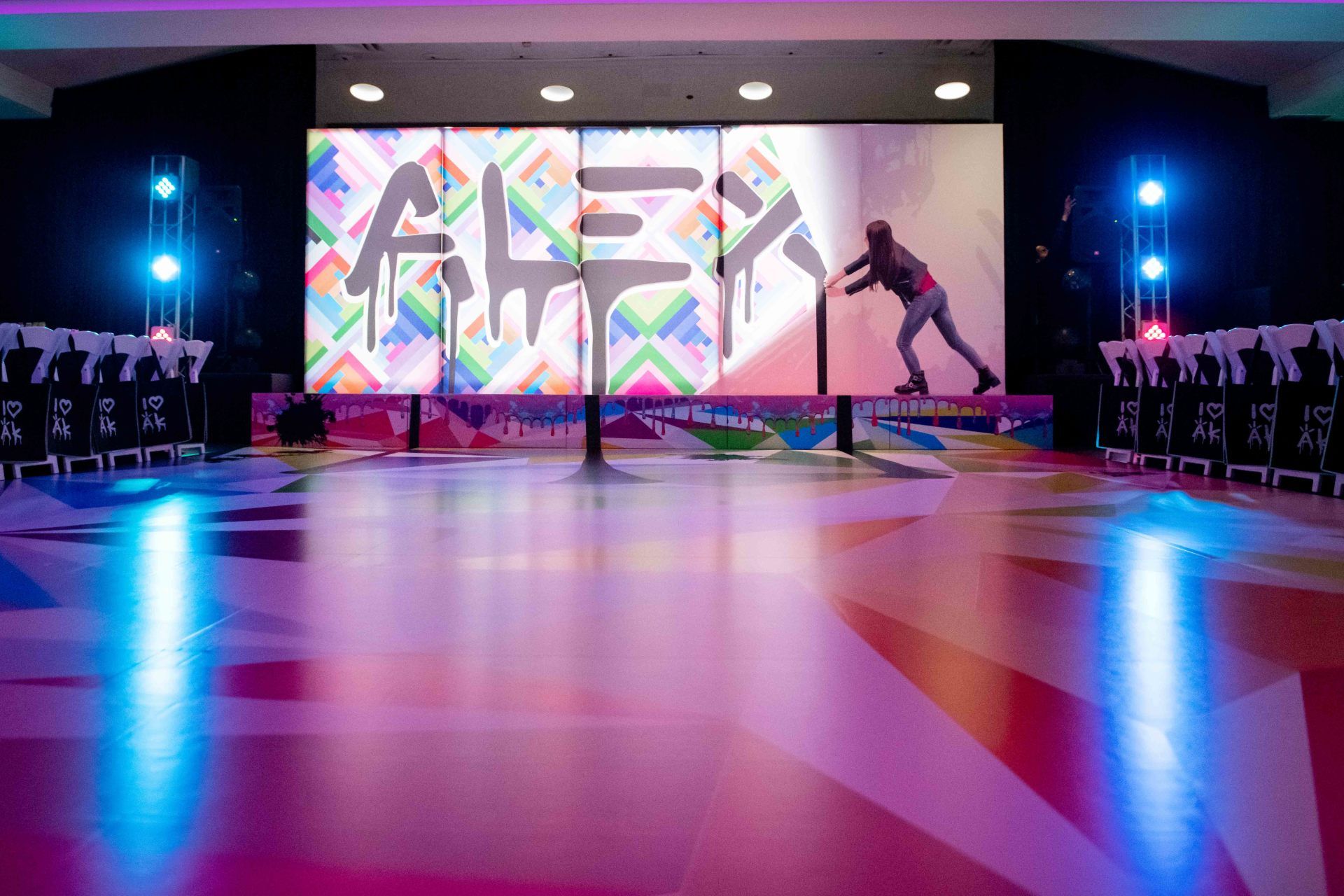 A man is skateboarding in front of a large screen that says alex