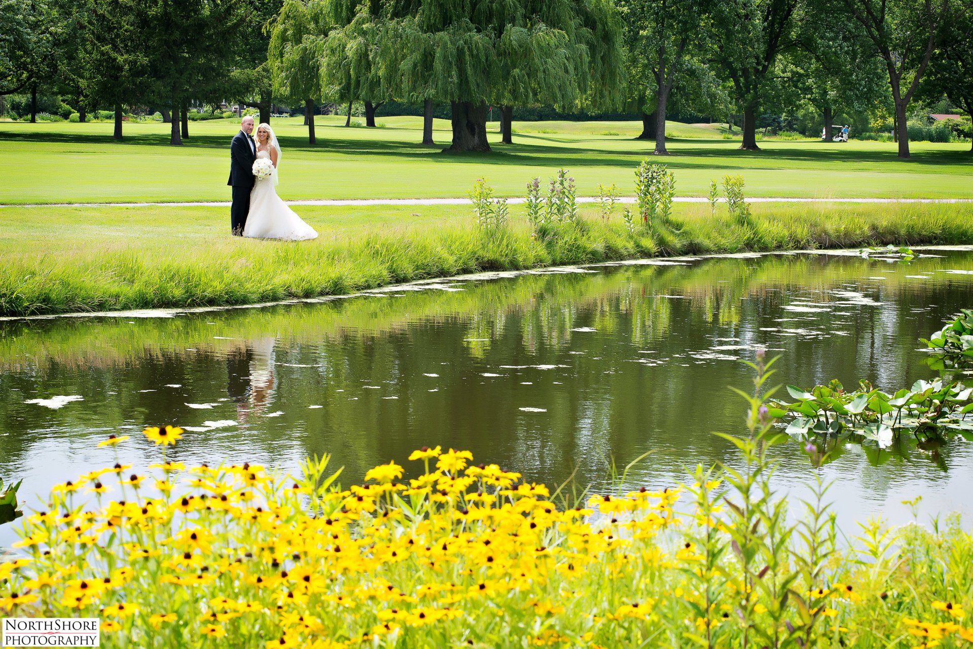 A bride and groom standing next to a pond in a park
