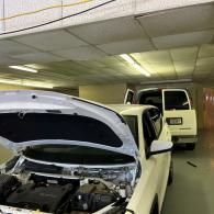 A white car with its hood open in a repair shop, with a white van parked behind it.