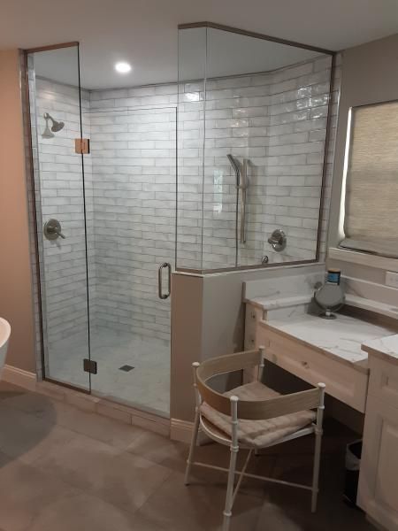 A modern walk-in shower with white brick-style tiles and a glass enclosure next to a bathroom vanity with a chair.