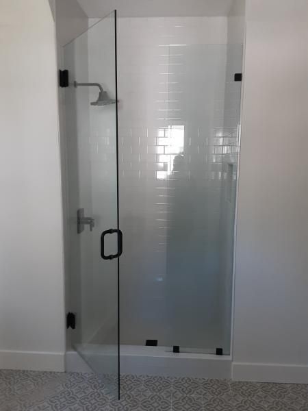 A glass walk-in shower with black metal hinges, a handle, and a shower head, set against a white-tiled wall.