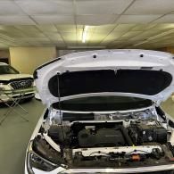 A white car parked indoors with its hood open, revealing the engine bay, with another car partially visible nearby.