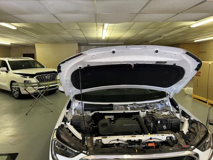 A white car with its hood open in a repair shop, with another white vehicle visible in the background.