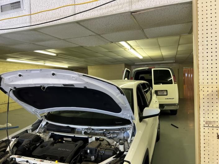 A white car with an open hood is parked inside a garage next to a white van.
